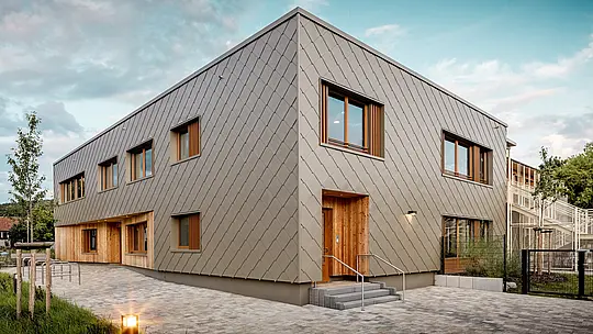 Sodobni vrtec v mestu Alzenau navdušuje s svojo jasno arhitekturo in visokokakovostno PREFA aluminijasto fasado v bronasti barvi. Fasada iz fasadnih rombov 44 × 44 daje stavbi eleganten, strukturiran videz in vizualno povezuje funkcionalne prostore v harmonično celoto. Lesene obloge na vhodih ustvarjajo topel, naraven kontrast z vzdržljivim aluminijem. Kombinacija masivne konstrukcije, lesenih elementov in obešene prezračevane fasade je primer trajnostnega gradbeništva. PREFA ovoj stavbe, ki ne zahteva veliko vzdrževanja, prepriča ne le s svojo reciklabilnostjo, ampak tudi s svojo odpornostjo proti vremenskim vplivom – idealno za prihodnostno usmerjene izobraževalne stavbe z visokimi arhitekturnimi zahtevami.