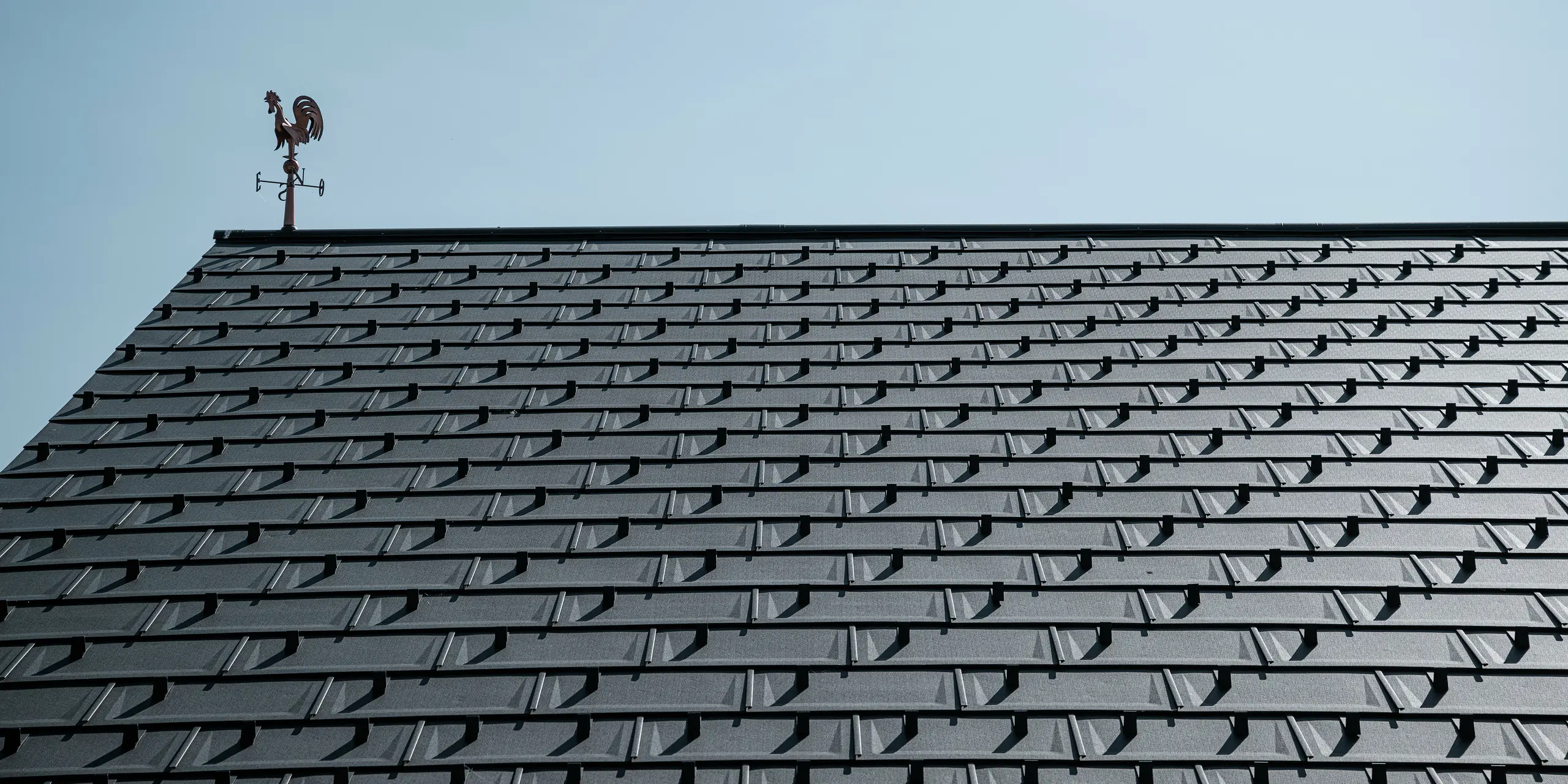Detailansicht eines PREFA Daches mit den Dachplatten R.16 in P.10 Funkelgrau. Die klare, moderne Optik der Aluminium-Dachplatten sorgt für eine langlebige, witterungsbeständige und wartungsfreie Dachlösung. Die Schneestopper bieten zusätzliche Sicherheit in schneereichen Regionen. Gekrönt wird das Dach von einer klassischen Wetterfahne, die Tradition mit moderner Technik verbindet. Dank der robusten PREFA Dachplatten R.16 bleibt das Dach über Jahrzehnte stabil und ästhetisch.