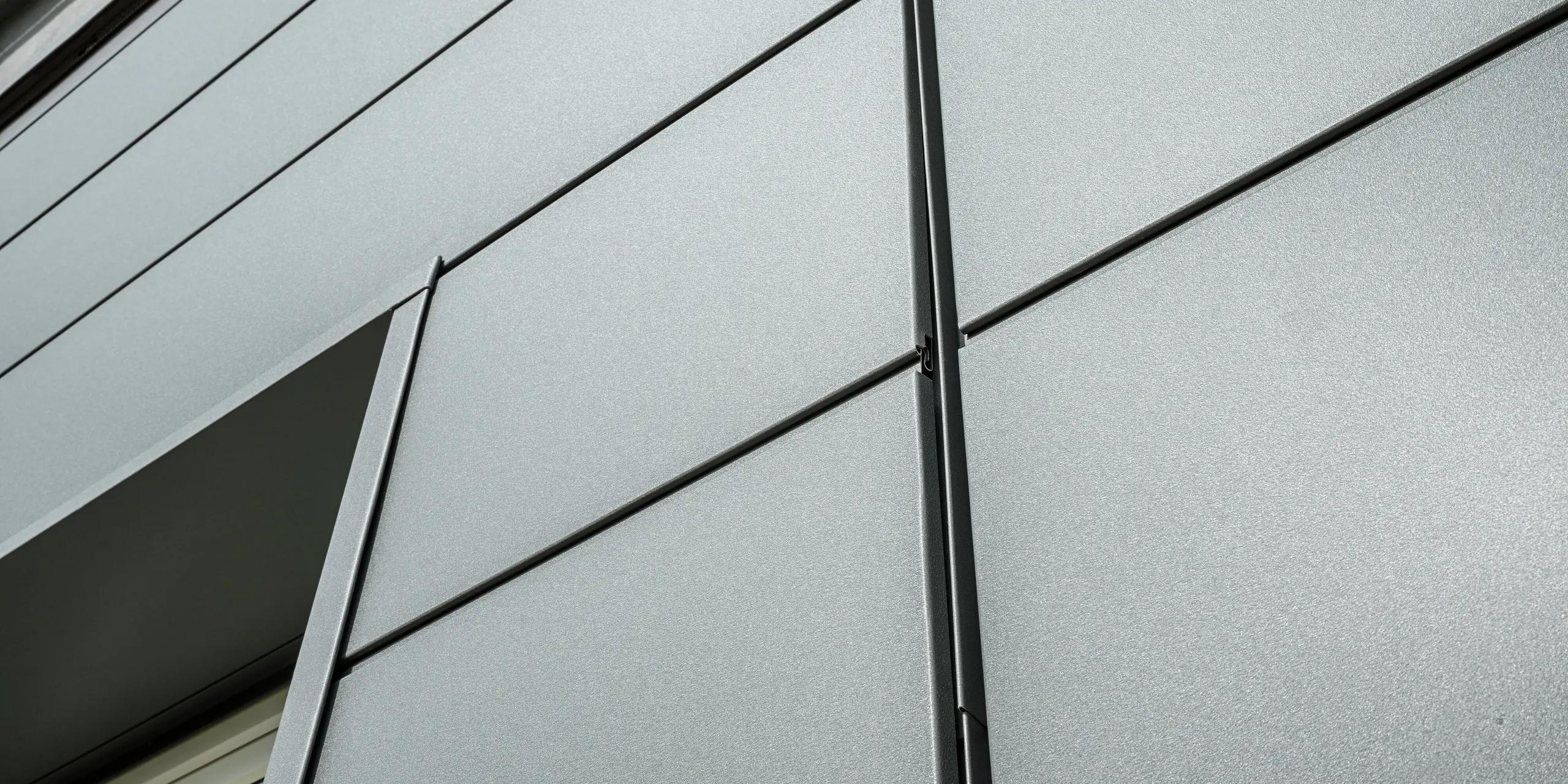 Die Fassade der Kaminkehrerinnung in Bindlach zeigt moderne PREFA Sidings in P.10 Dunkelgrau. Die klar strukturierten Aluminium-Paneele sind nicht nur ein optisches Highlight, sondern auch äußerst widerstandsfähig gegenüber Witterungseinflüssen. In der Detailaufnahme sind die präzise verlegten Blech-Paneele und die stilvolle, schlichte Linienführung deutlich zu erkennen.