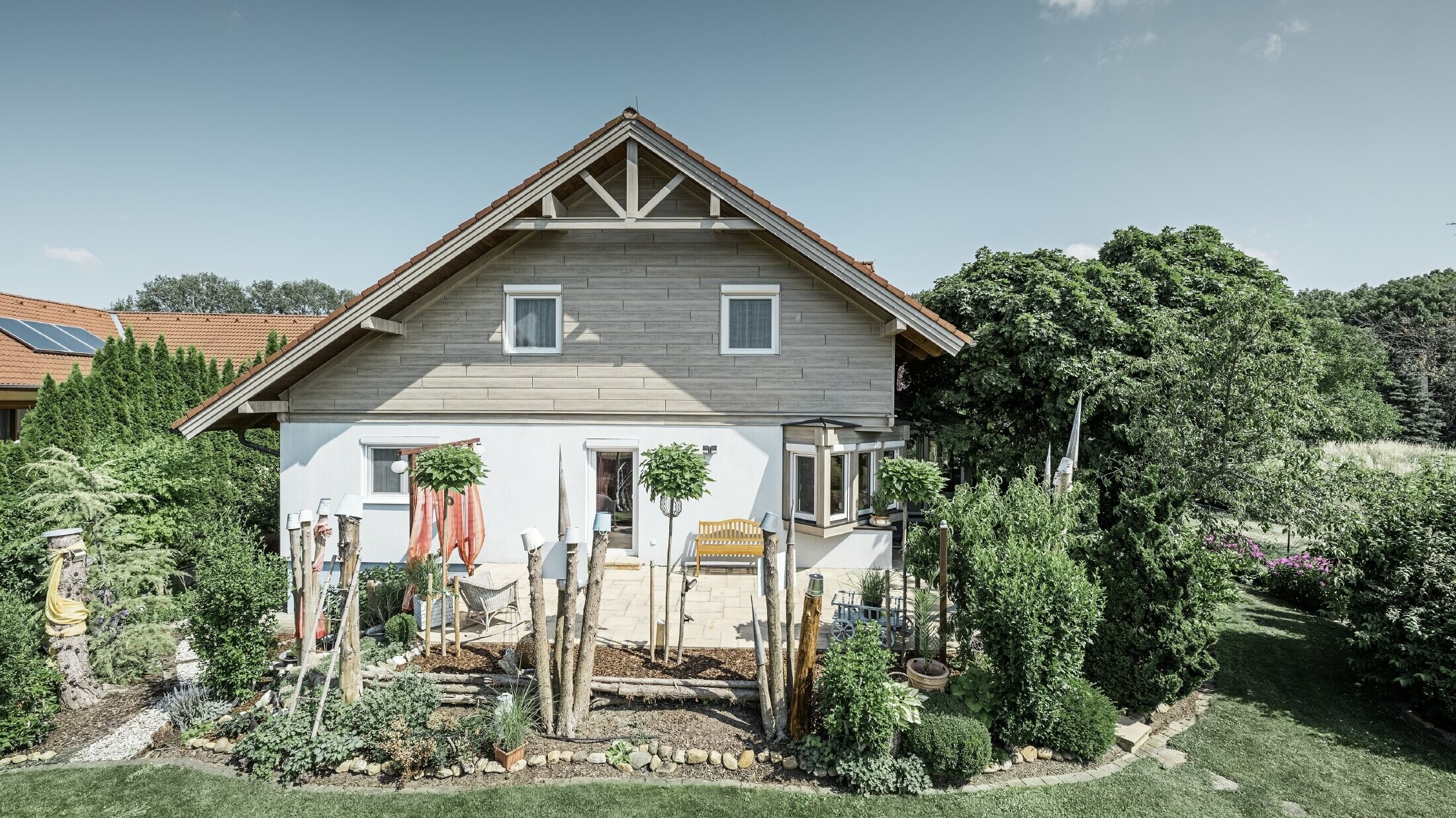 Enodružinska hiša z obnovljeno fasado in prenovljenim oblogom čela v sodobnem lesnem videzu v sivo-bežni barvi, v kombinaciji z ometano fasado, dvokapno streho in urejenim vrtom.