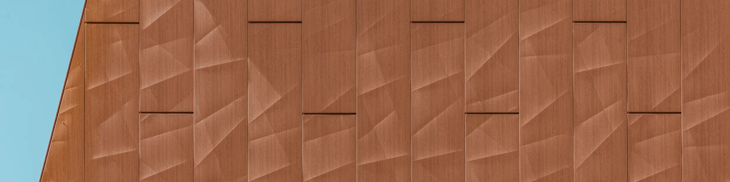 Detajlni posnetek aluminijaste fasade **PREFA Siding.X** na črpalni postaji za pitno vodo WML v Lottumu z natančno zgibanimi paneli v topli rjasto-rdeči (rjavi) optiki kot vizualnim poudarkom.  Geometrično strukturirana površina ustvarja fascinantno igro svetlobe, senc in finih linij, ki se glede na zorni kot dinamično spreminja.  Visokokakovostni aluminijasti paneli PREFA prepričajo z majhno težo, visoko odpornostjo na vremenske vplive, barvno obstojnostjo in popolno možnostjo recikliranja – za trajno, trajnostno in skoraj brez vzdrževanja fasadno rešitev.