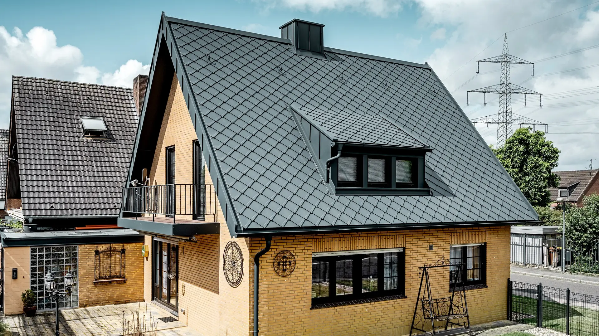 Enodružinska hiša iz rumene opeke po obnovi strehe s PREFA strešnimi ploščicami strešni romb 29 × 29 v temno sivi barvi; novo pokrita, strma dvokapna streha z rombastim vzorcem, vgrajenim podstrešjem in dimnikom, teraso in vrtom v ospredju.