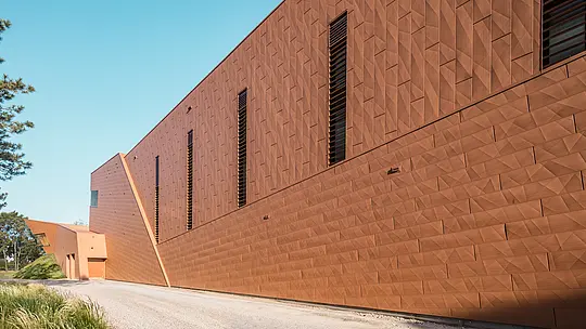 Dinamična fasada črpalne postaje za pitno vodo WML v Lottumu z natančno razporejenimi, ozkimi okenskimi odprtinami, ki pritegnejo pogled. Izrazita aluminijasta fasada PREFA Siding.X v topli rjavi barvi ustvarja živahno igro svetlobe, sence in geometrije vzdolž jasne robne linije stavbe. Izjemna struktura združuje tehnično funkcionalnost s skulpturalno estetiko. Lahki, trajni in v celoti reciklirani PREFA aluminijasti paneli prepričajo z visoko odpornostjo proti vremenskim vplivom, oblikovno stabilnostjo ter trajnostno, vzdrževanju prijazno ovojnico stavbe.