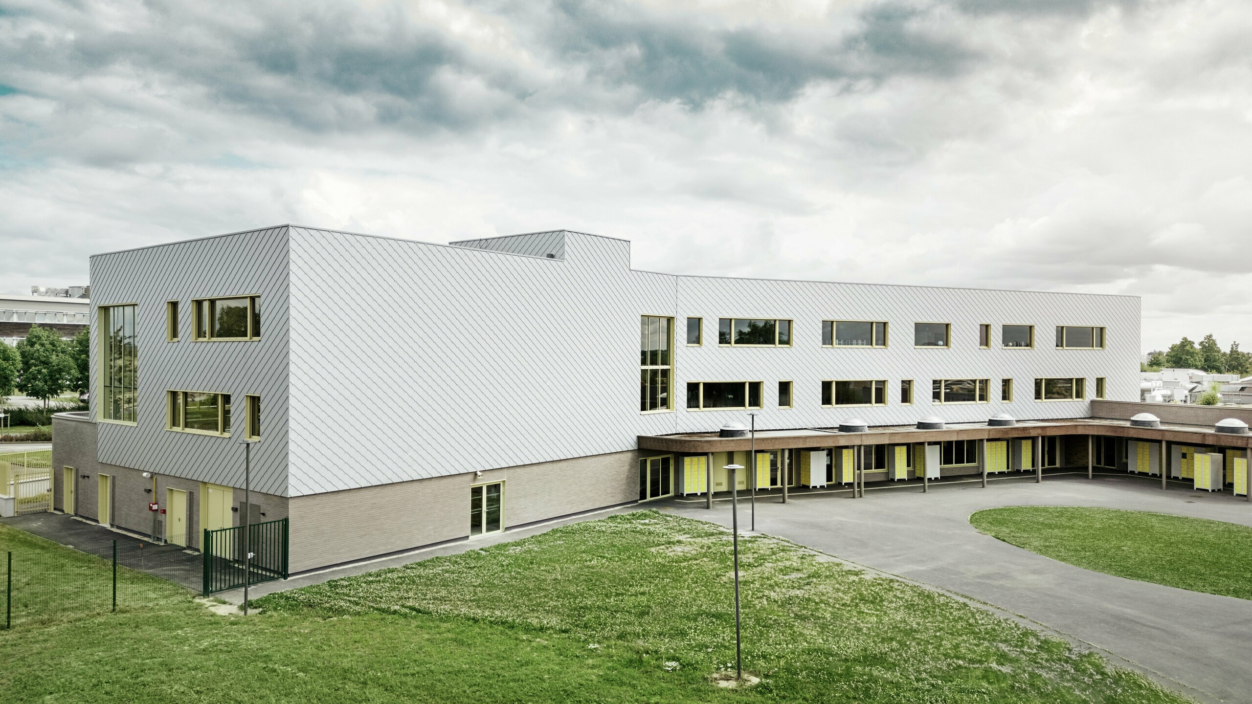 Slika prikazuje zadnjo stran šole Collège Léontine Dolivet. V ospredju je prostrano, asfaltirano šolsko dvorišče z jasnimi linijami in odprtimi površinami, ki se lahko uporabljajo za odmore in prostočasne dejavnosti. Nekaj zelenih površin popestri okolico. V ozadju se dviga moderna šolska stavba z izrazito aluminijasto fasado iz kovinsko srebrnih PREFA fasadnih rombov, ki odbijajo svetlobo in ustvarjajo dinamičen učinek. Več velikih oken zagotavlja svetlost v notranjosti. Slika je posneta pri dnevni svetlobi, ob rahlo oblačnem nebu, kar še posebej poudarja kovinsko površino.