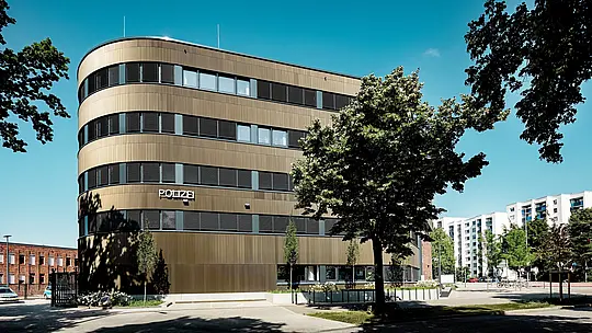 Dinamičen arhitekturni posnetek policijske postaje 43 v Hamburgu: ukrivljena nova stavba s PREFA aluminijasto fasado iz ekstrudiranih zobčastih profilov v barvi srednje bronaste privlači vse poglede kot izrazit poudarek v mestni podobi. Fino strukturirana površina ustvarja živahne svetlobne in senčne učinke med vodoravnimi pasovi oken ter poudarja tekoče linije fasade. PREFA aluminij prepriča z visoko odpornostjo proti koroziji, dolgo življenjsko dobo in majhnimi stroški vzdrževanja – idealno za trajnostne, funkcionalne in arhitekturno natančne fasadne rešitve.