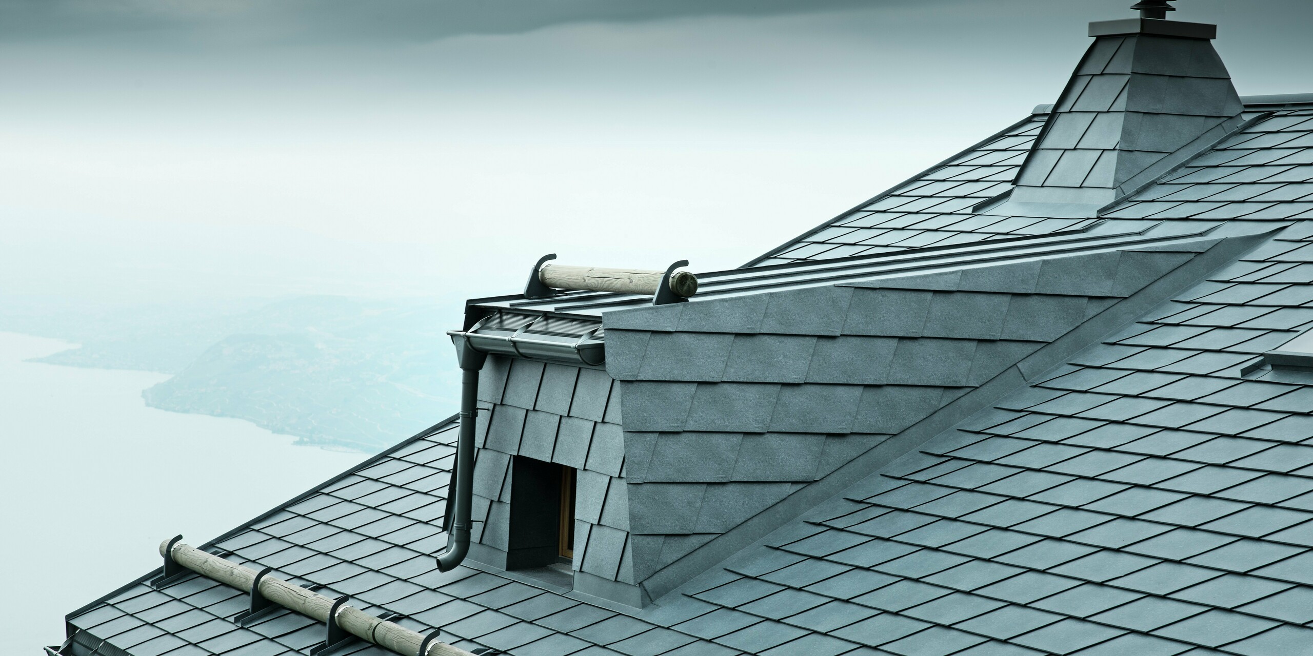 Die Dachansicht dieser modernen Holz-Lodge in der Schweiz zeigt steingraue PREFA Aluminium-Dachschindeln und eine große Dachgaube. Das langlebige Aluminiumdach schützt das Gebäude vor den extremen Wetterbedingungen am Genfersee. Der raffinierte Schneefang aus Rundholz und der markante Kamin betonen das einzigartige Design der Architektur.