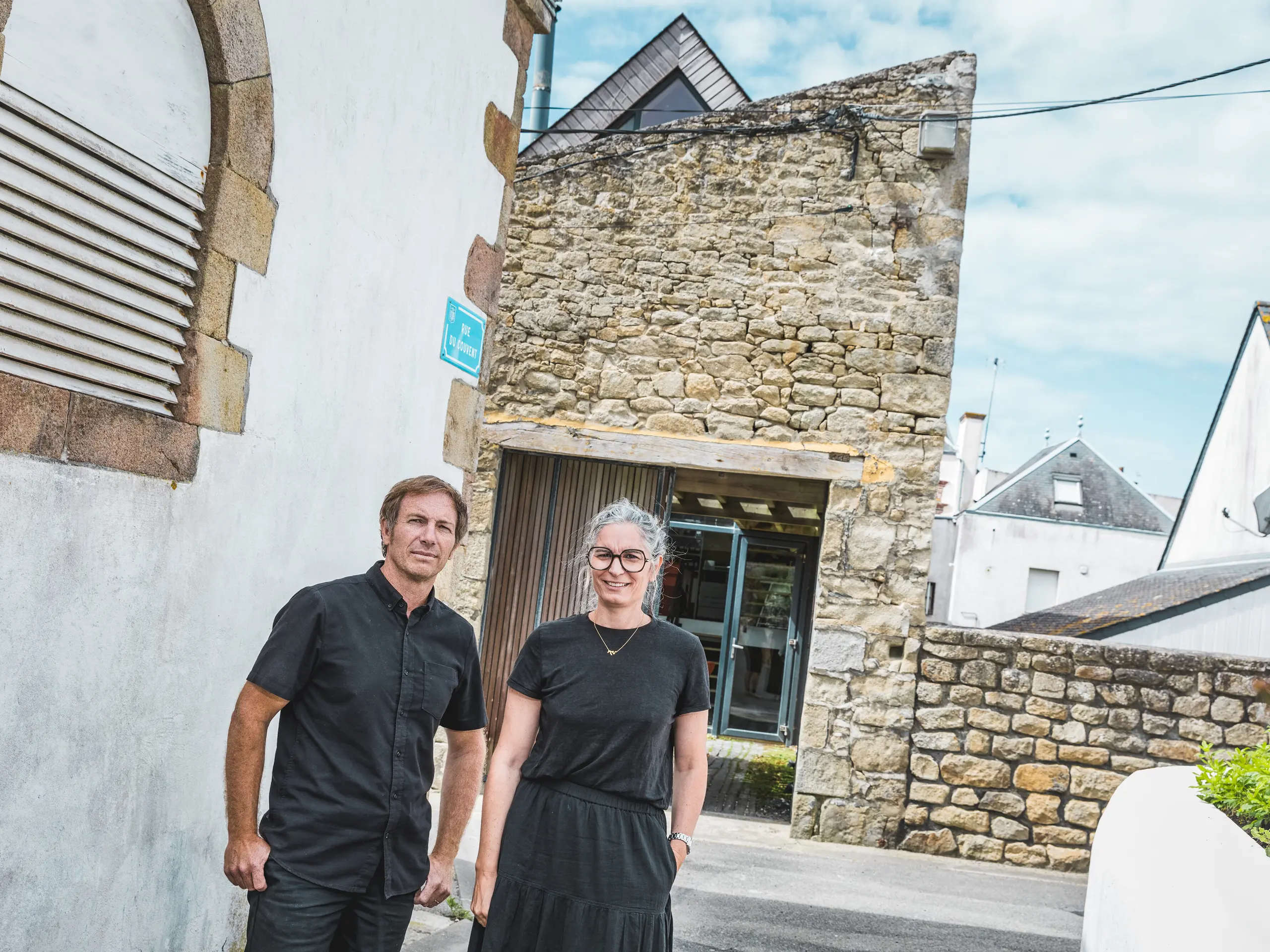 V ozki bretonski ulici stojita arhitekta Anne-Charlotte in Jean-François Riguidel pred izrazito fasado iz naravnega kamna. Oba sta oblečena v črno. Zanimiv kontrast med tradicionalnim zidovjem, belima fasadama hiš in sodobno zasnovo streh usmerja pogled na uspešno povezovanje regionalne gradbene tradicije in sodobnega oblikovanja.