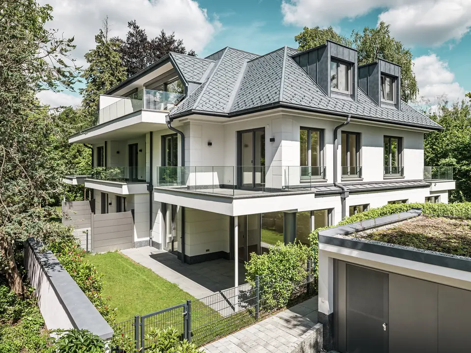 Ansicht eines modernen Mehrfamilienhauses in der Nähe von München mit stilvollem Design. Das Dach wurde mit PREFA Dachrauten 29 × 29 in P.10 Steingrau gedeckt. Die markante Dachkonstruktion wird durch elegante Flachdachgauben aus PREFALZ akzentuiert, die sowohl ästhetisch als auch funktional überzeugen. Der gepflegte Gartenbereich sowie die großzügige Terrasse betonen die hochwertige Architektur und schaffen eine harmonische Verbindung zwischen Innen- und Außenbereich. Die langlebigen und witterungsbeständigen PREFA-Produkte garantieren eine perfekte Integration in die grüne Umgebung und bieten dauerhaft Schutz und Schönheit.