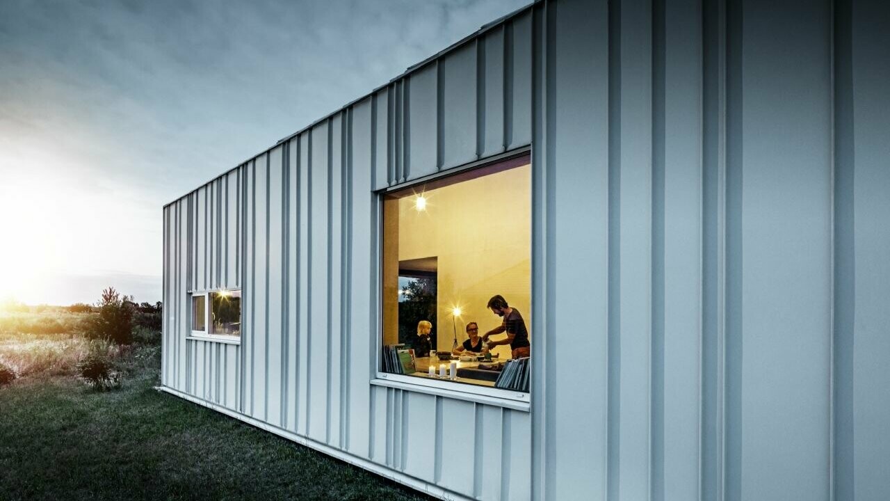 Fasada s pravokotnim pokončnim zgibom PREFALZ z različnimi širinami trakov v barvi P.10 prefa bela, fotografirano v mraku. Skozi jedilnico vidimo družino pri večerji.