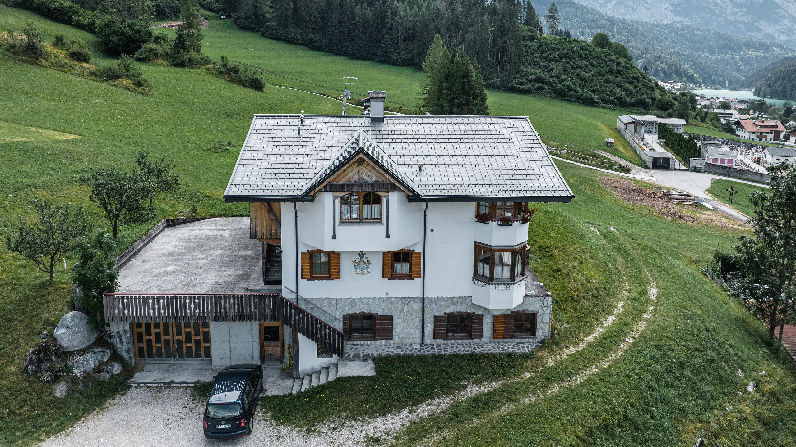 Pogled s ptičje perspektive na vhod v tradicionalno planinsko kočo v Auronzo di Cadore po prenovi strehe z aluminijastimi strešniki PREFA v svetlo sivi barvi P.10. Stavba z značilnim alpskim šarmom in sodobno pločevinasto streho se harmonično zliva z bujno zeleno pokrajino in impresivno gorsko panoramo v ozadju ter kaže na uspešno kombinacijo funkcionalnosti in regionalne arhitekture.