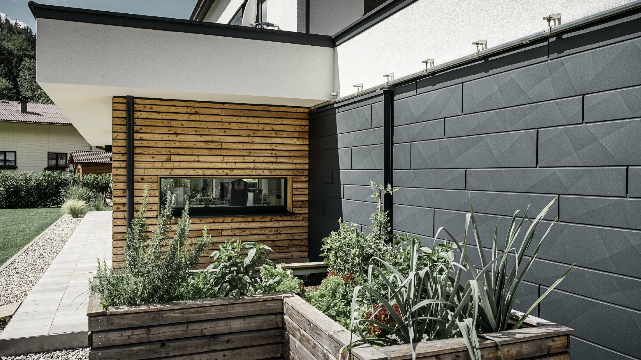 Ta fotografija prikazuje bližnji posnetek enodružinske hiše, kjer je v ospredju fasadna obloga iz PREFA Siding.X v barvi P.10 antracitna. Temni fasadni paneli z izrazitim gubastim videzom ustvarjajo privlačen vizualni kontrast z živahnimi, z lesom obloženimi deli in okoliškimi zelenimi površinami. Pred fasado iz aluminija so postavljene visoke grede, zasajene z različnimi rastlinami in grmičevjem, kar poudarja naraven in trajnosten videz hiše. Kombinacija sodobnih materialov in vključitev v vrtno okolje ustvarja podobo sodobne arhitekture, ki se harmonično vključuje v svoje okolje.