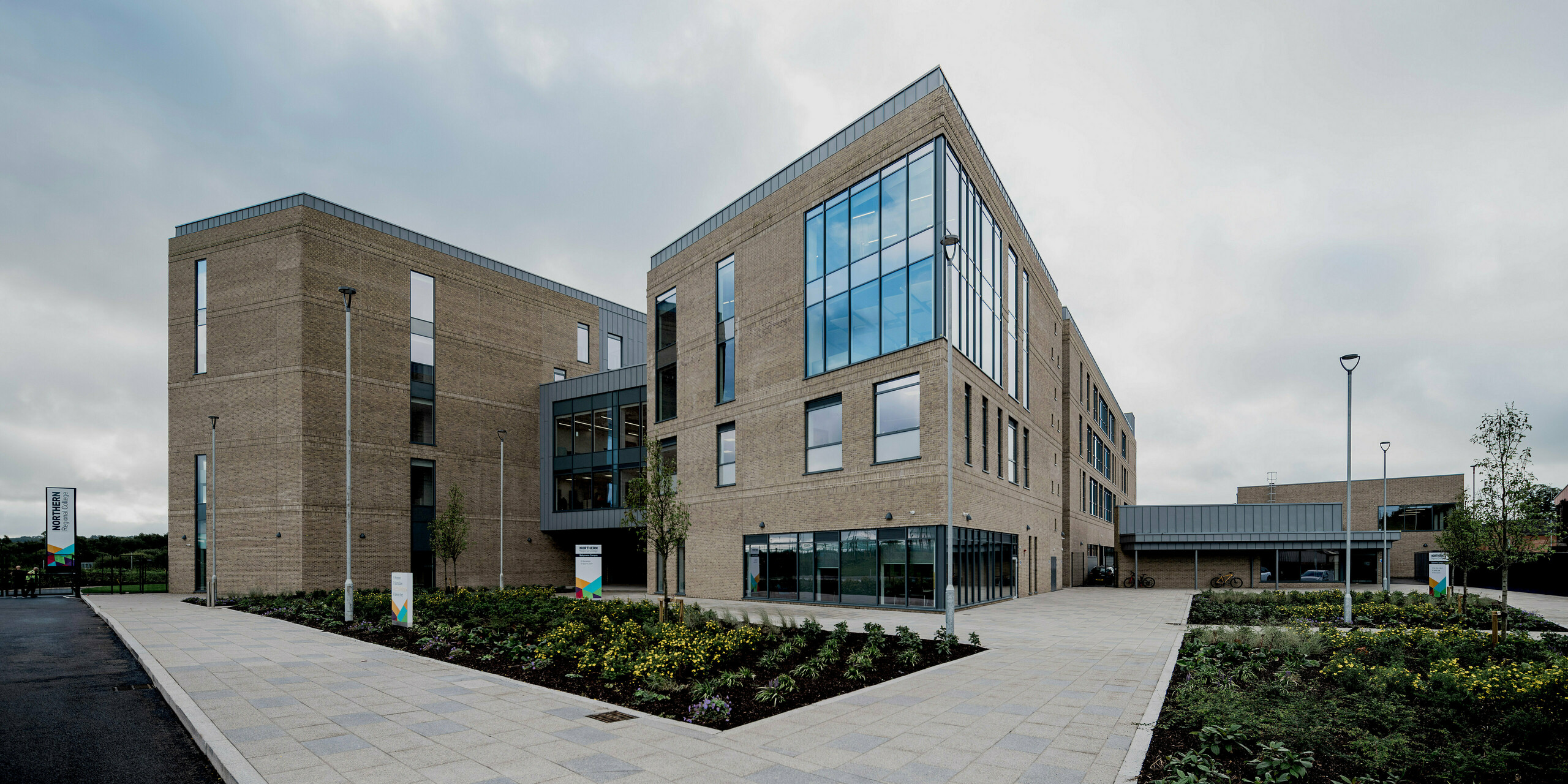 Eine großzügige Gesamtansicht des Northern Regional College in Ballymena zeigt mehrere klar gegliederte Baukörper aus hellem Klinker, verbunden durch Glasflächen und offene Wege. Entlang der Dachkanten und Fassadenabschlüsse setzen PREFALZ Aluminium-Stehfalzbahnen in Patinagrau präzise horizontale Akzente. Die Metallflächen rahmen die Architektur elegant und kontrastieren mit dem Mauerwerk und dem weiten Himmel. PREFA Aluminium überzeugt durch Langlebigkeit, Wartungsarmut und eine zeitlos moderne Gestaltung für Bildungsbauten.