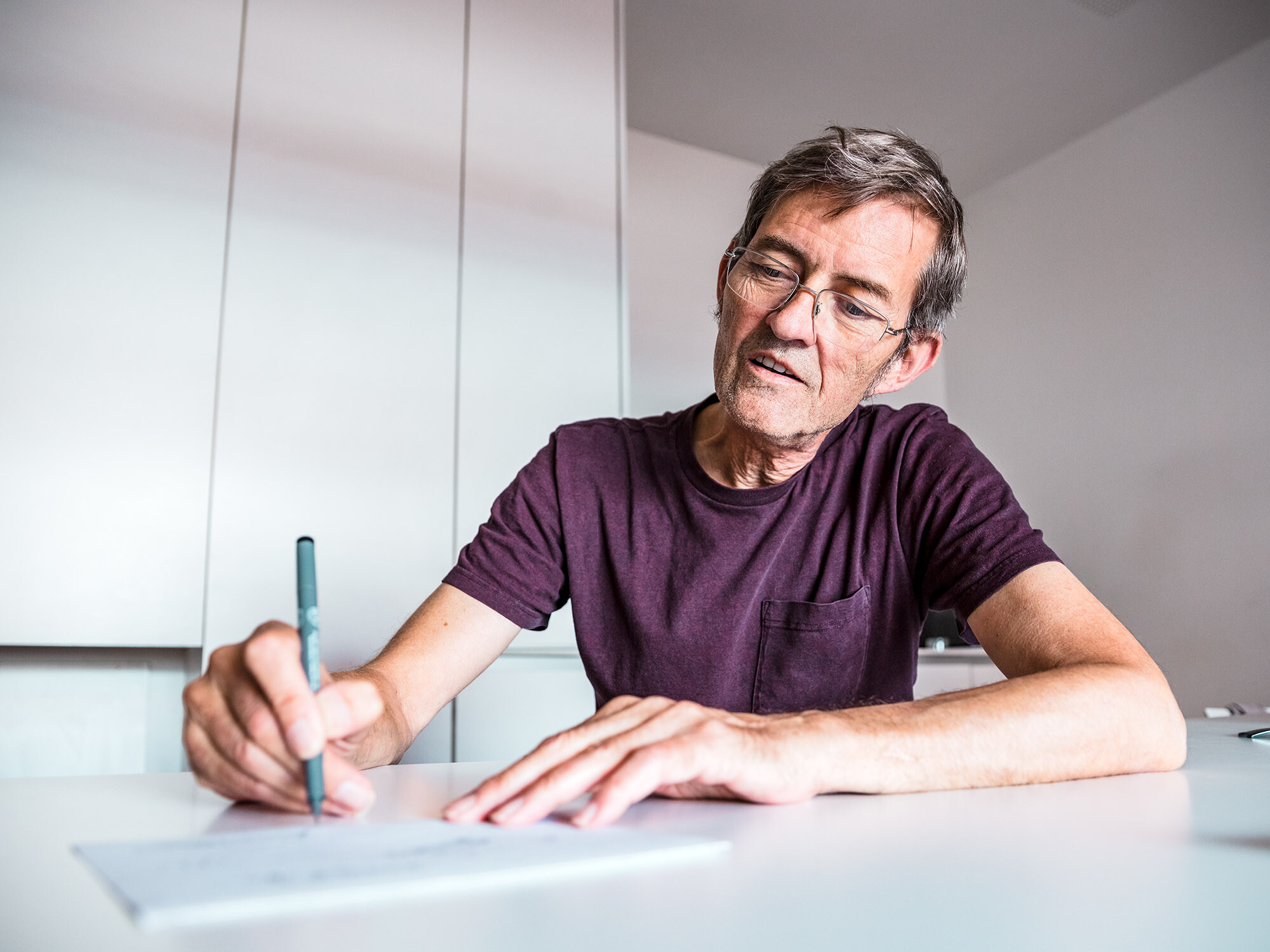Architekt Dominique Cornaert sitzt an einem weißen Tisch und zeichnet, hinter ihm sind weiße Wände sowie ein weißer Schrank zu sehen.