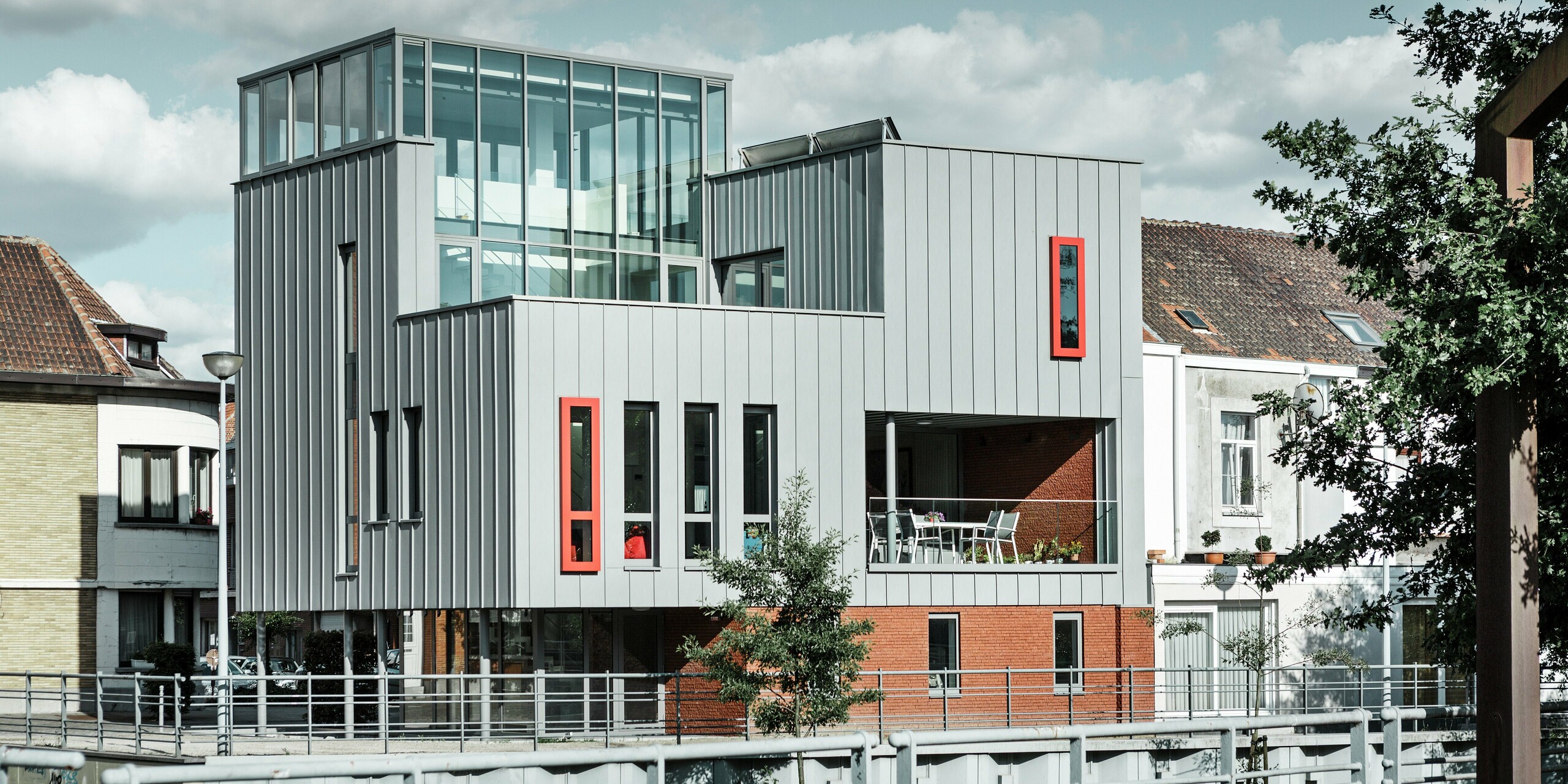 [Translate to Slovenščina:] Moderne Architektur am Flussufer: Das Wohn-und Bürogebäude kombiniert eine elegante PREFALZ Fassade in Patinagrau mit auffälligen, roten Fensterrahmen als Eyecatcher. Die klar strukturierte Stehfalzoptik trifft auf großzügige Glasflächen und eine überdachte Terrasse. Aluminium als Baumaterial überzeugt hier durch Wetterbeständigkeit, Formstabilität und zeitloses Design.