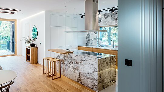 Die Aufnahme zeigt die elegante Innenarchitektur der Budapester Mehrgenerationen‑Villa mit einer modernen, offen gestalteten Küche. Markante Elemente sind die marmorierten Natursteinoberflächen der Kücheninsel und Rückwand, kombiniert mit warmen Eichenholzakzenten und hochwertigen Einbaumöbeln. Großzügige Fensterflächen lassen viel Tageslicht herein und verbinden den Raum harmonisch mit dem umliegenden Grün. Die klare Linienführung, die edle Materialwahl und die präzise Ausführung spiegeln den hochwertigen architektonischen Anspruch des Gebäudes wider, dessen äußere Gestaltung durch langlebige PREFA Aluminiumprodukte ergänzt wird.