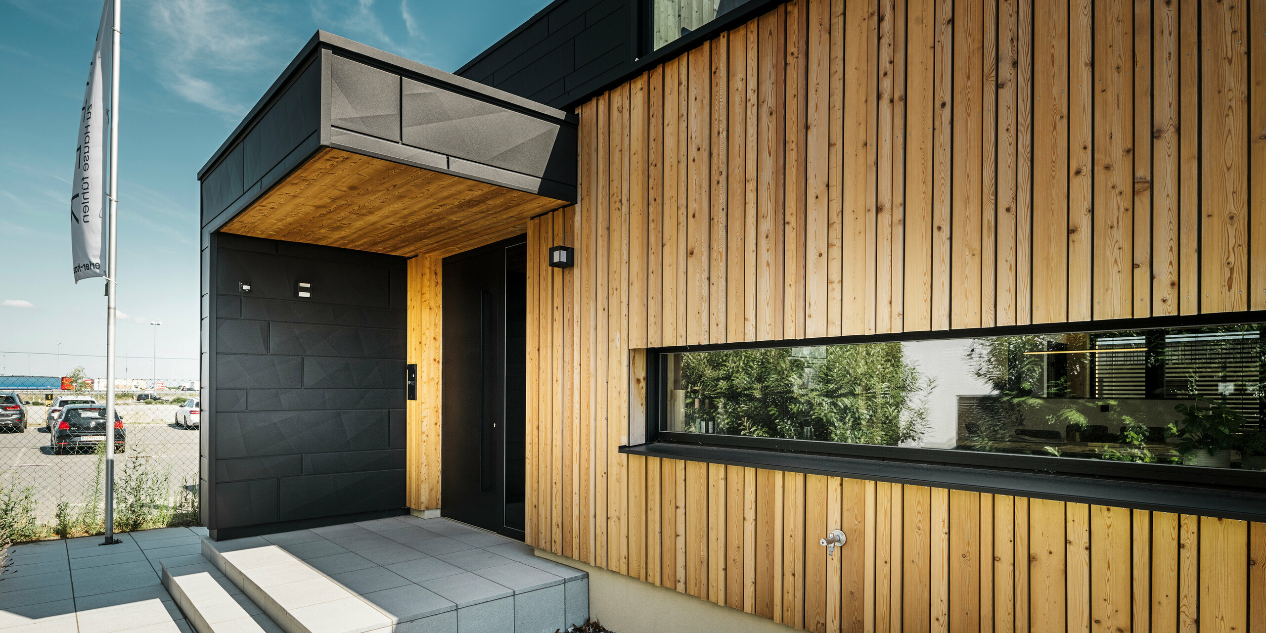 Stranski vhod v vzorčno hišo Autark v centru montažnih hiš Blaue Lagune v Vösendorfu s sodobno fasado iz naravnega lesa in črnih plošč PREFA Siding.X. Slika prikazuje elegantno kombinacijo lesene obloge in aluminija v visokokakovostnem premazu P.10 črni barvi.