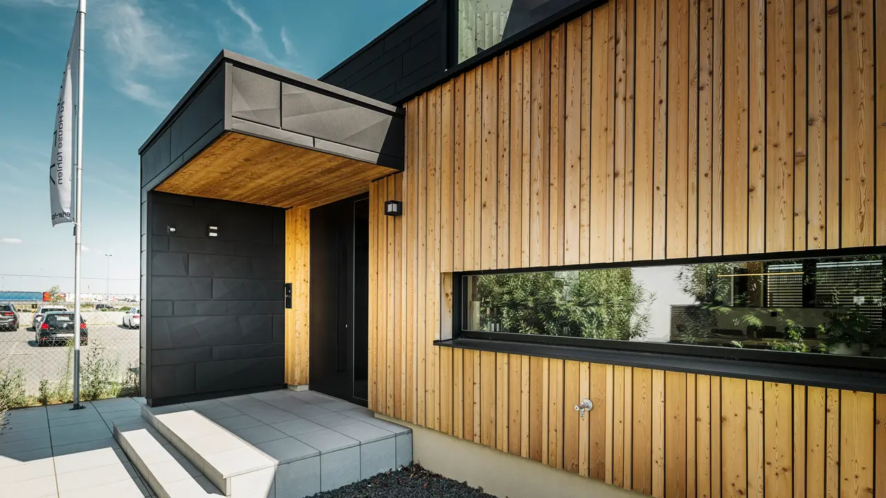 Stranski vhod v vzorčno hišo Autark v centru montažnih hiš Blaue Lagune v Vösendorfu s sodobno fasado iz naravnega lesa in črnih plošč PREFA Siding.X. Slika prikazuje elegantno kombinacijo lesene obloge in aluminija v visokokakovostnem premazu P.10 črni barvi.