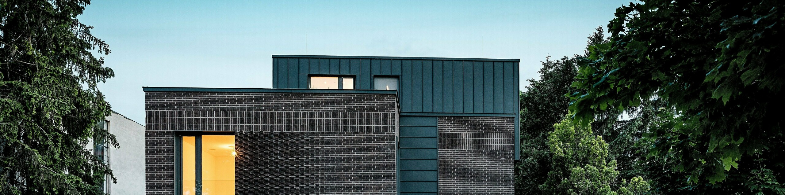 Na sliki je prikazana sodobna večstanovanjska stavba v Budimpešti, katere kubična strešna konstrukcija iz PREFALZ aluminija v barvi P.10 antracit tvori izrazit kontrast z zgodovinskim okoljem. Vertikalni kotni stoječi zgibi ustvarjajo zanimivo igro svetlobe in poudarjajo jasen arhitekturni jezik. Visokokakovostni PREFA aluminijasti izdelki stavbi ne dajejo le elegantnega, brezčasnega videza, temveč prepričajo tudi s svojo dolgo življenjsko dobo, odpornostjo proti vremenskim vplivom in natančno izdelavo. Antracitno obarvana fasada in streha delujeta kot močan magnet za poglede in poudarjata inovativen značaj stavbe.