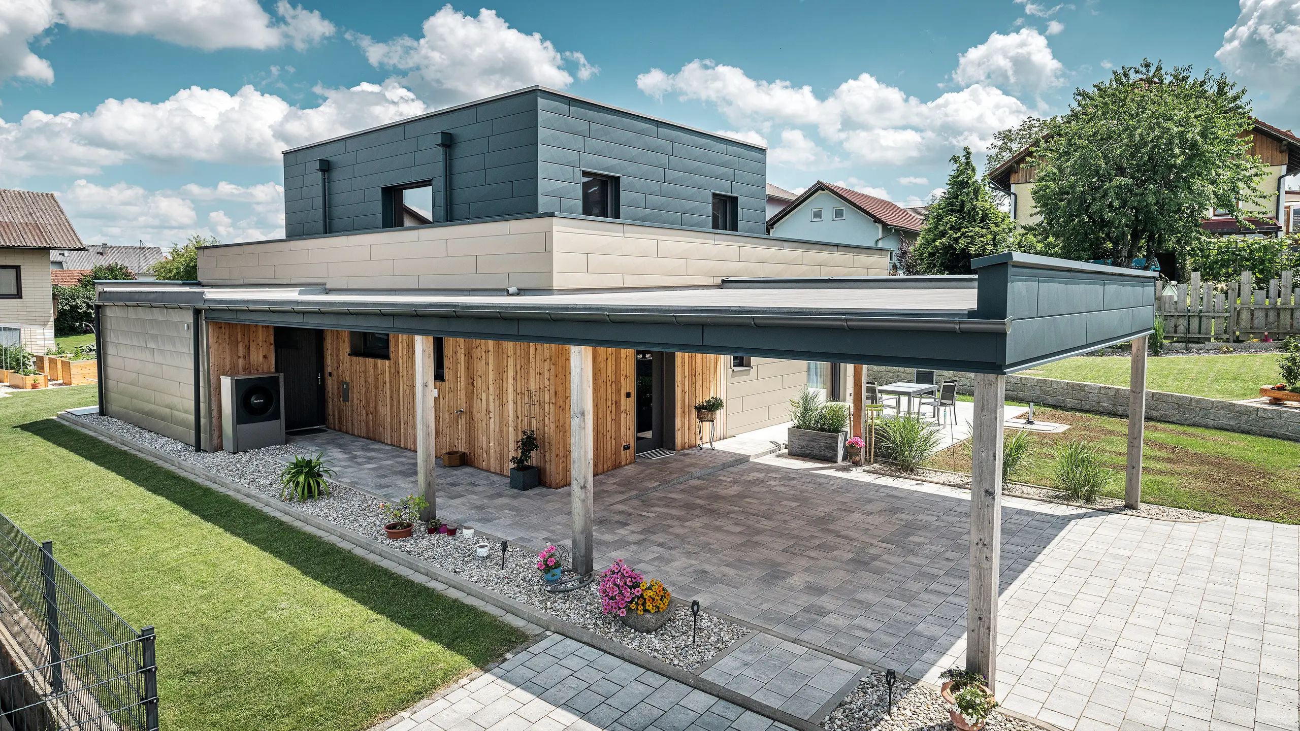 Letalski posnetek dostopa do moderne enodružinske hiše v Wippenhamu v Avstriji z dvobarvno PREFA aluminijasto fasado v bronasti in antracitni barvi. Hiša prikazuje stilsko integracijo fasadnih panelov Siding.X in lesene obloge. Jasno vidna je pokrita garaža, ki je brezhibno integrirana v arhitekturo hiše. Urejena vrt in terasa poudarjata sodoben in funkcionalen dizajn stavbe.
