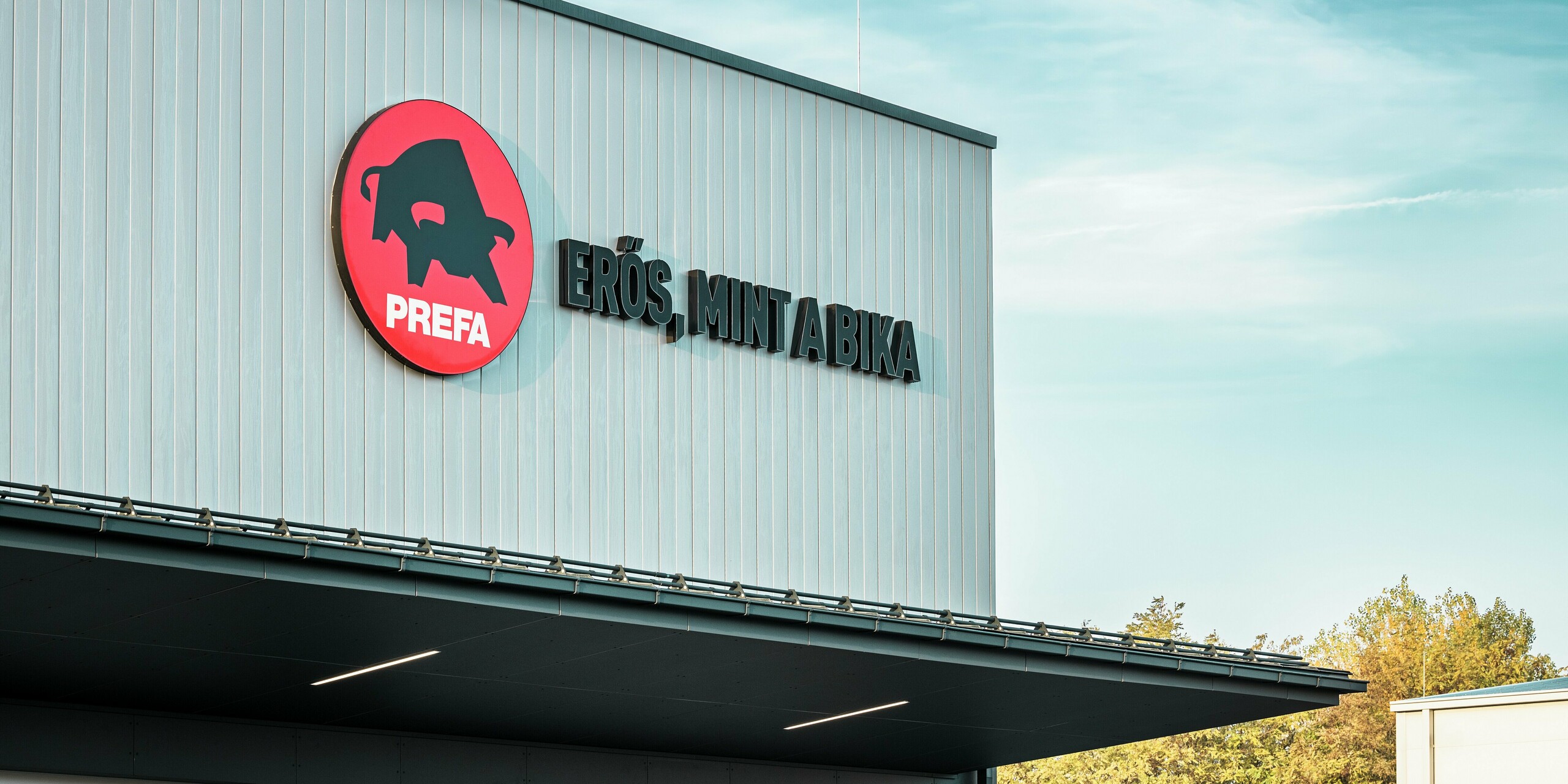 Prepoznavni logotip PREFA in madžarski slogan »Erős, mint a bika« (»Močan kot bik«) zaznamujeta fasado PREFA lokacije v Törökbálintu. Obloge s PREFA elementi Siding v srebrno kovinski barvi poskrbijo za moderen videz, vrhunsko odpornost na vremenske vplive in dolgo življenjsko dobo. Natančna izvedba poudarja visok standard kakovosti podjetja PREFA in odraža moč blagovne znamke.