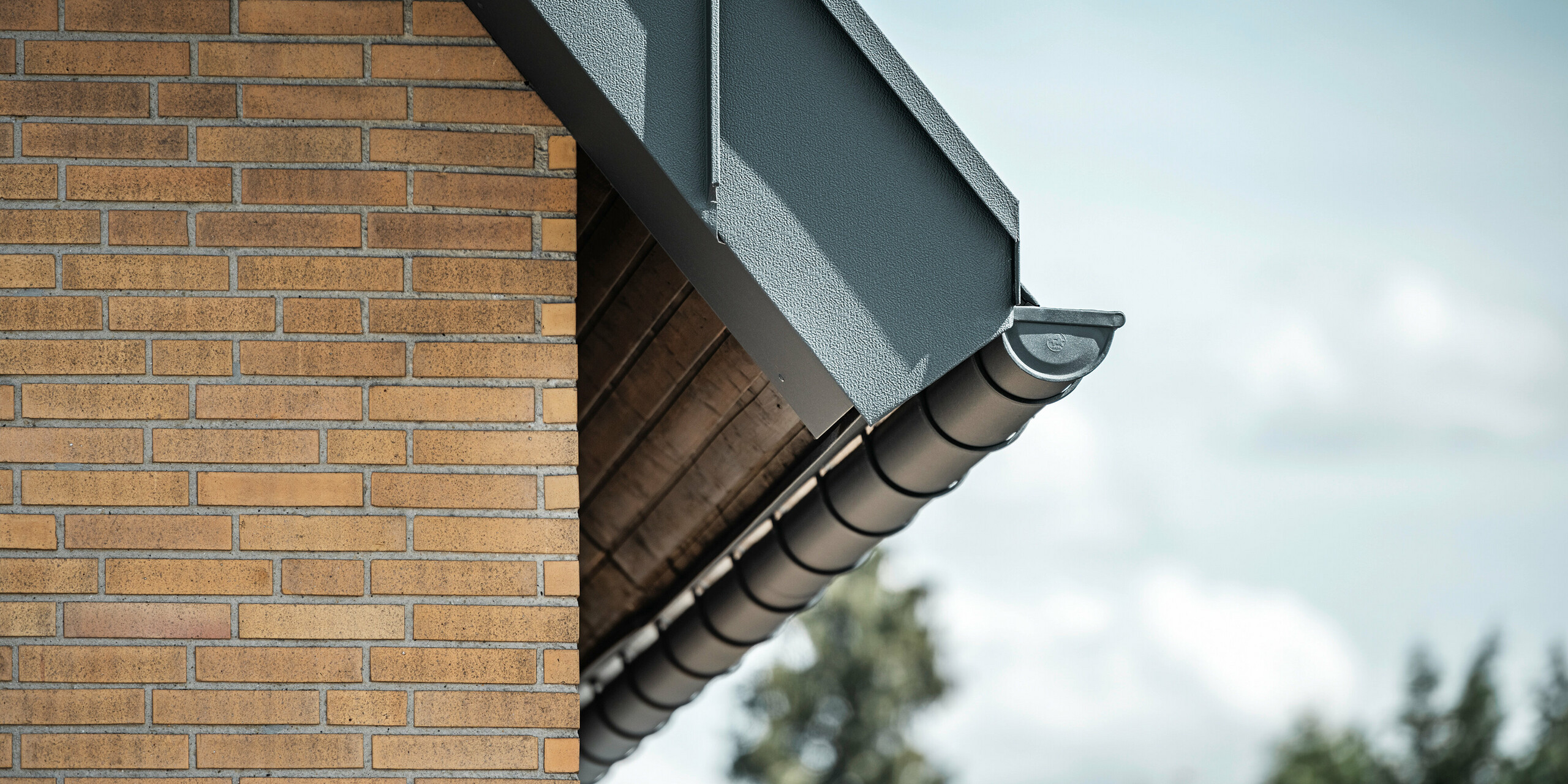 Podroben prikaz PREFA sistema za odvodnjavanje strehe v antracitni barvi. Žleb jasno izstopa iz toplih tonov opečne stene in poudarja natančno izdelavo in kakovost uporabljenih materialov.