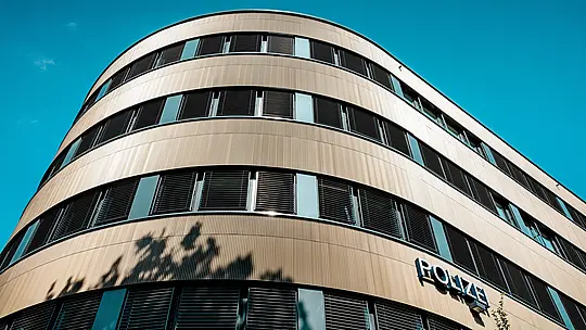 Dramatičen pogled od spodaj na ukrivljeno novo stavbo policijske postaje 43 v Hamburgu: izrazita PREFA aluminijasta fasada iz eloksiranih ekstrudiranih zobčastih profilov v barvi srednje bronaste postane arhitekturni magnet za poglede. Fino rebrasta površina ustvarja živahne svetlobne odseve in poudarja tekoče zaobljenosti ter vodoravne pasove oken. Zahvaljujoč visoki odpornosti proti koroziji, dolgoživosti in minimalnim zahtevam po vzdrževanju rešitve iz aluminija PREFA ponujajo trajnostno, funkcionalno in estetsko zahtevno oblikovanje fasade.