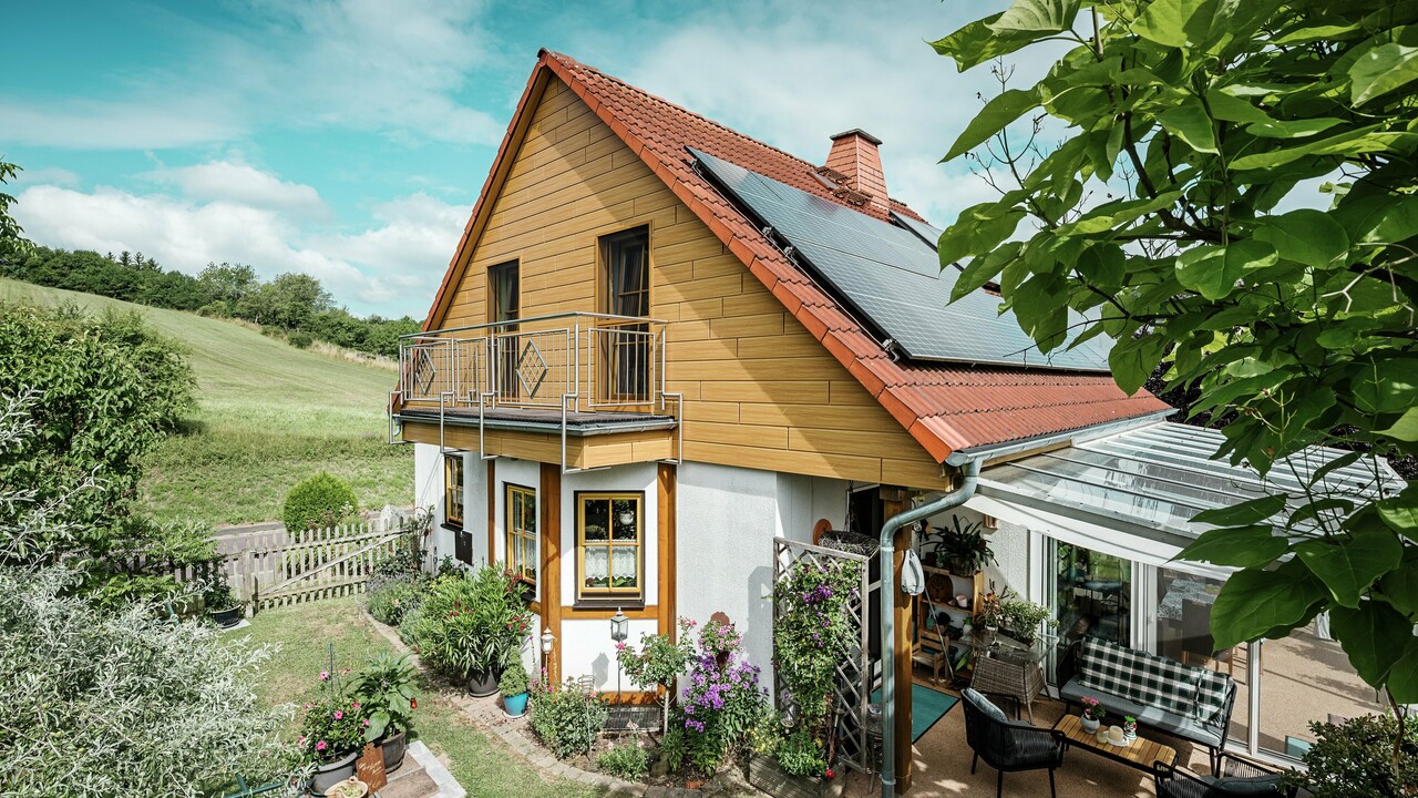 Ta slika prikazuje očarljivo enodružinsko hišo v Blieskastlu, katere sveže prenovljen zatrep s paneli PREFA Siding v odtenku naravni hrast takoj pritegne pogled. Topel videz lesa iz trpežnega aluminija se izjemno skladno ujema z rdečo streho iz strešnikov in prostornim zimskim vrtom. Hišo obdajajo bujno zelenje in skrbno urejene cvetlične gredice, kar ustvarja idiličen in živahen prizor.