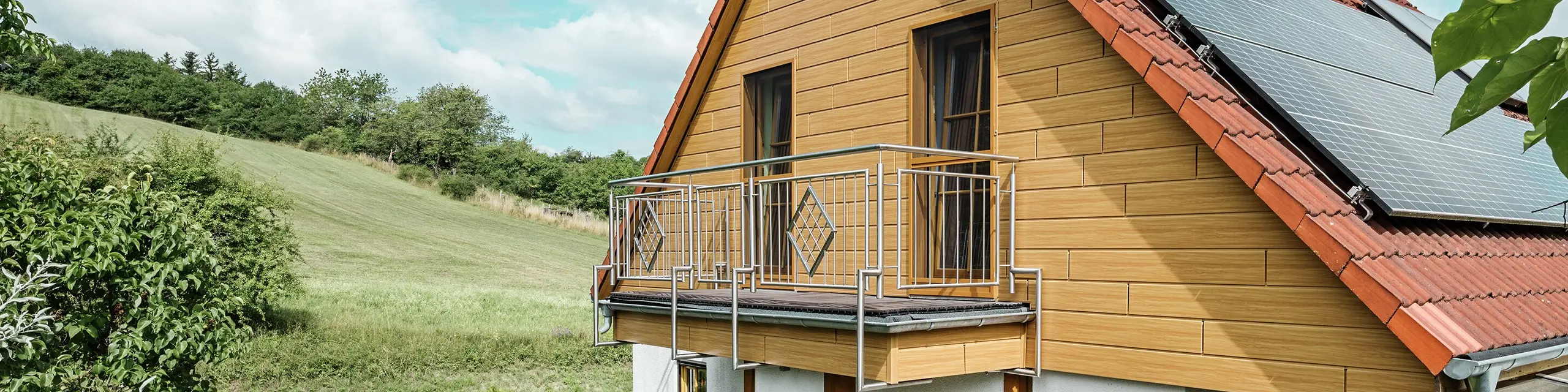 Ta slika prikazuje očarljivo enodružinsko hišo v Blieskastlu, katere sveže prenovljen zatrep s paneli PREFA Siding v odtenku naravni hrast takoj pritegne pogled. Topel videz lesa iz trpežnega aluminija se izjemno skladno ujema z rdečo streho iz strešnikov in prostornim zimskim vrtom. Hišo obdajajo bujno zelenje in skrbno urejene cvetlične gredice, kar ustvarja idiličen in živahen prizor.