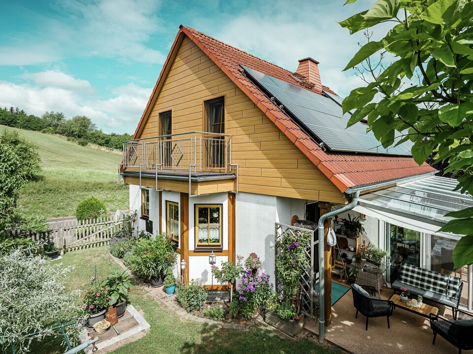 Dieses Bild zeigt ein charmantes Einfamilienhaus in Blieskastel, dessen frisch sanierter Giebel mit PREFA Sidings in Eiche Natur sofort ins Auge fällt. Die warme Holzoptik aus langlebigem Aluminium harmoniert beeindruckend mit dem roten Ziegeldach und dem großzügigen Wintergarten. Umgeben von üppigem Grün und liebevoll angelegten Blumenbeeten entsteht eine idyllische, lebendige Szene. Die robuste PREFA Fassade verleiht dem Haus nicht nur einen natürlichen Look, sondern überzeugt auch durch Witterungsbeständigkeit, Farbtreue und nahezu wartungsfreie Langlebigkeit.