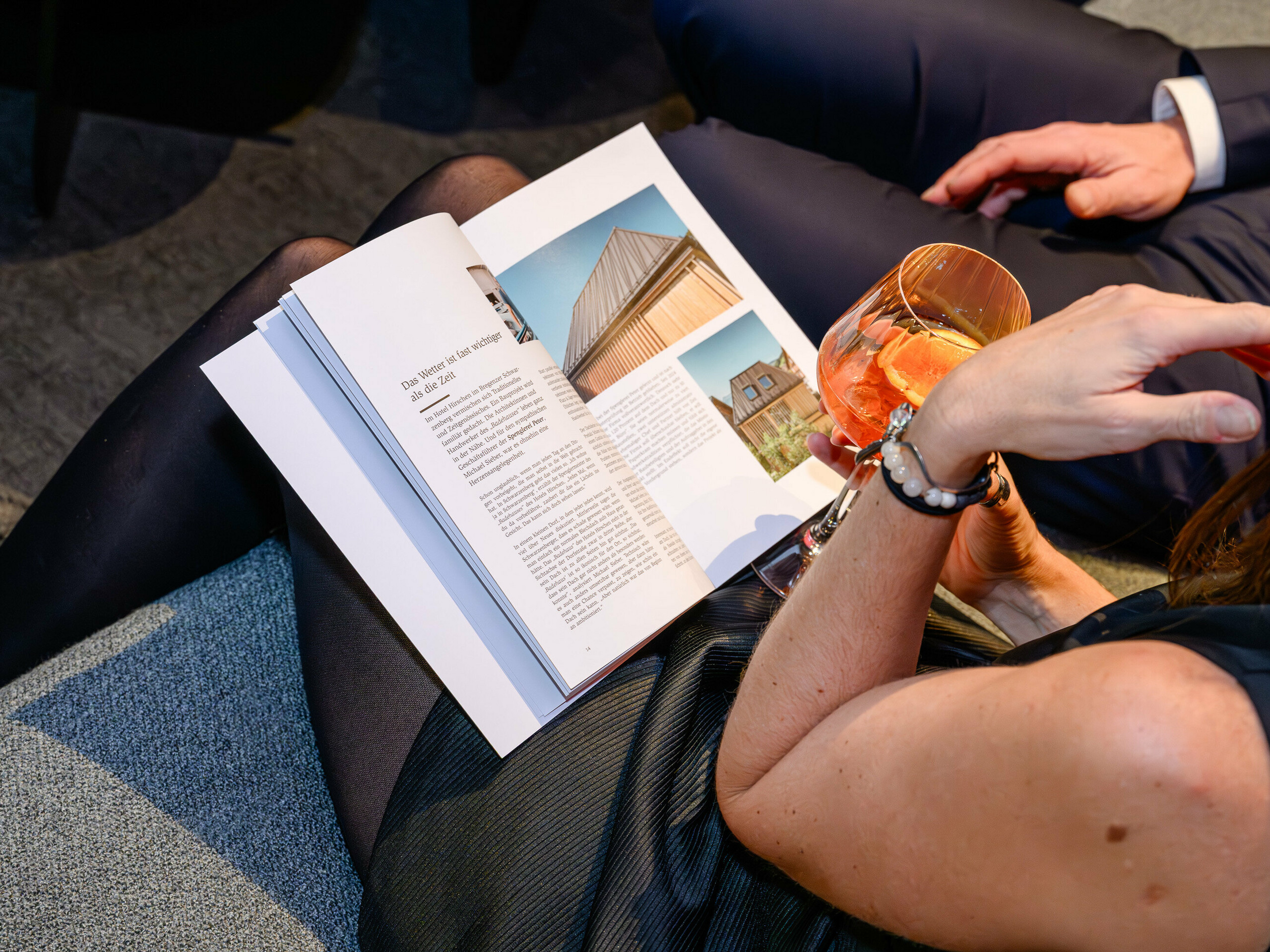 Učinkovita scena na predstavitvi PREFARENZEN 2026 v k47.wien: elegantna ženska s pijačo v roki listuje novo knjigo o arhitekturi PREFA – privlačen izdelek, poln navdiha za oblikovanje. Vidna je stavba s sistemom PREFALZ, ki navdušuje s sodobnim aluminijastim videzom. Slika izraža užitek, izmenjavo in radovednost do inovacij. Visokokakovostna PREFA površina združuje estetiko in trajnost – idealna za kreativne arhitekturne ideje. Aluminij PREFA postavlja merila in navdušuje tako strokovnjake kot načrtovalce, arhitekte in obdelovalce.