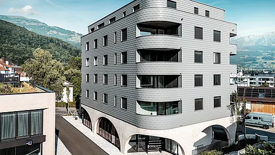 Zunanji pogled na hišo Bernerhaus v Buchsu z značilno trikotno obliko in nežno zaobljenimi balkonskimi pasovi. Fasada iz PREFA fasadnih skodel v barvi P.10 svetlo siva obdaja stavbo kot luskasta aluminijasta koža in ustvarja natančen, živahen videz fasade. Nad masivnim betonskim podstavkom z elegantnimi arkadami deluje lahka, vremensko odporna PREFA aluminijasta fasada še posebej izrazito. Trajna, enostavna za vzdrževanje in oblikovno stabilna postavlja pred alpskim gorskim ozadjem sodoben arhitekturni poudarek.
