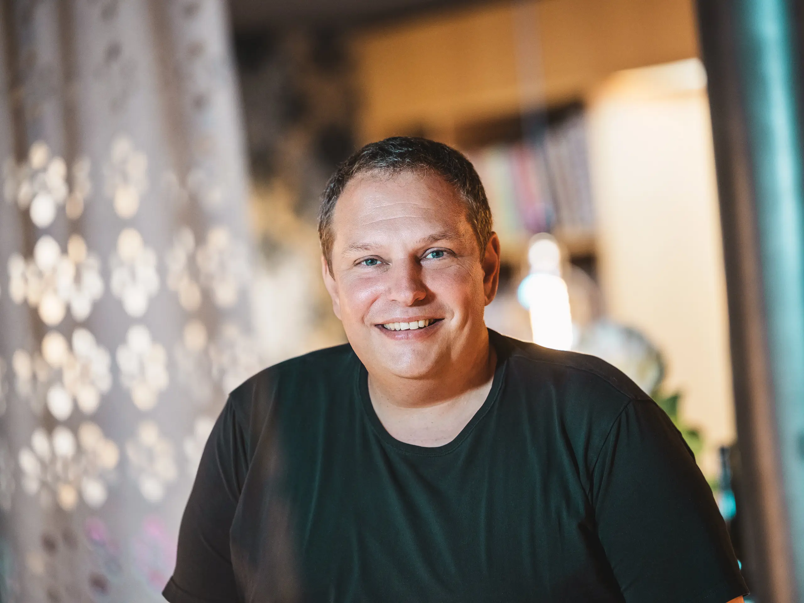 Portret vorarlberskega arhitekta Jürgena Hallerja, ki je zasnoval hišo Immi v Ebnitu kot dosledno minimalistično in trajnostno leseno hišo. Nasmejani arhitekt stoji v toplo osvetljenem notranjem prostoru. Njegov projekt združuje regionalne materiale z robustno PREFA solarno streho iz solarnih strešnih plošč in strešnih plošč R.16 v barvi P.10 črna, prilagojeno alpskim razmeram.
