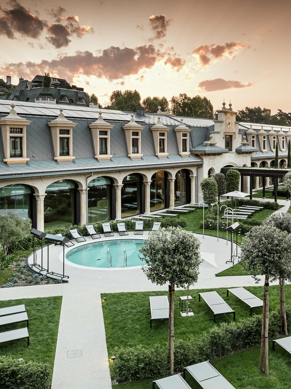 Abendstimmung im Thermalpark „De Montel – Terme Milano“ mit eindrucksvollem Blick auf die denkmalgeschützte Fassadenarchitektur im Jugendstil. Die Dacheindeckung mit PREFA Dachrauten 44 × 44 in P.10 Hellgrau verbindet Nachhaltigkeit mit Ästhetik. Im Vordergrund laden Liegewiesen, gepflegte Wege, Olivenbäume und ein rundes Thermalbecken zum Verweilen ein.