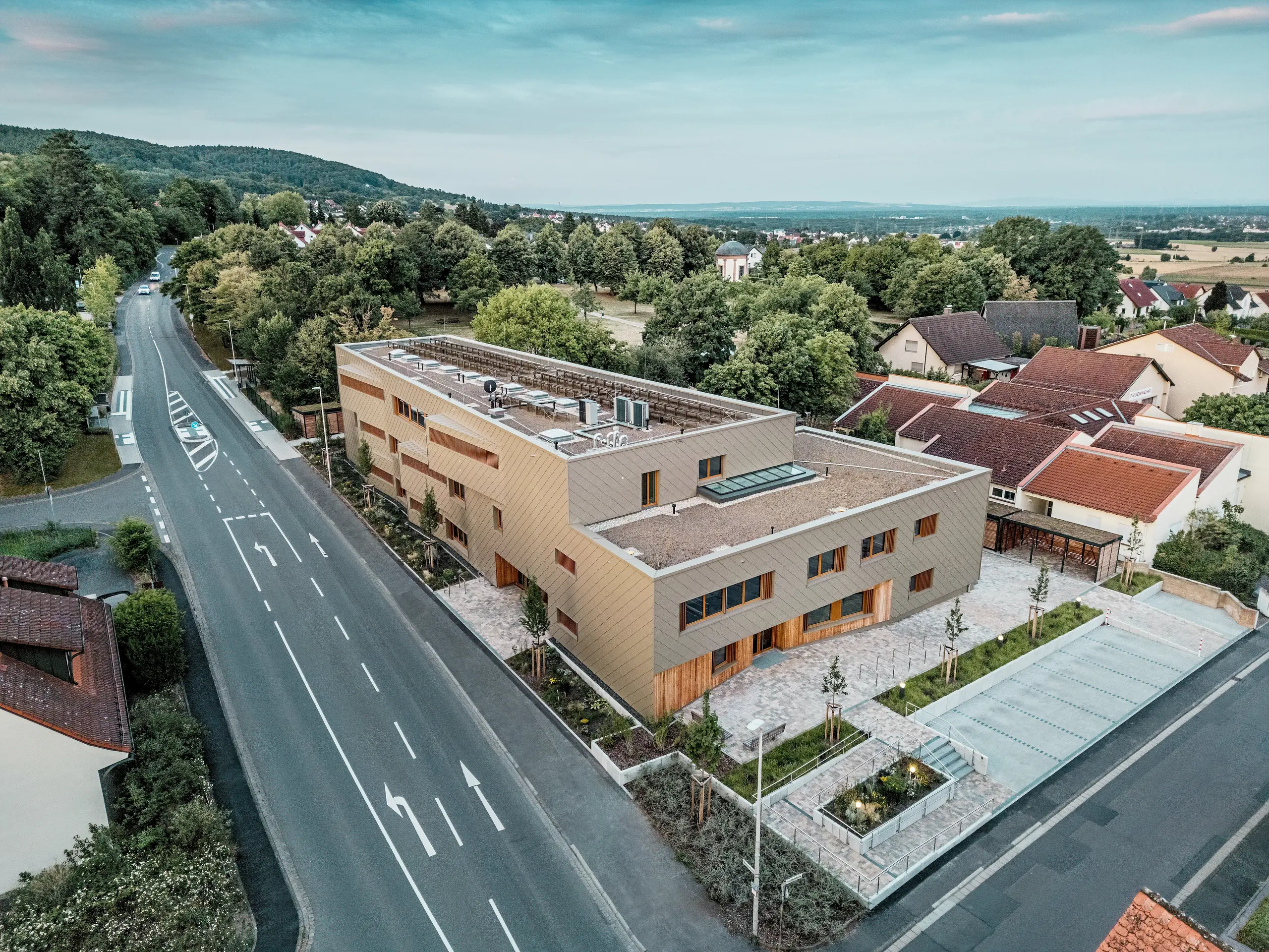 Impresivna celostna zasnova občinskega vrtca v Alzenau je vidna na letalski fotografiji. Kotna hibridna stavba je harmonično vključena v mirno stanovanjsko četrt na robu Spessarta. Fasada iz bronastih PREFA fasadnih rombov 44 × 44 daje stavbi izrazit, visokokakovosten videz in poudarja moderno arhitekturo. Zeleni strešni površini, prostrani zunanji prostori in jasna ureditev prometa poudarjajo trajnostni celostni koncept. Kombinacija trajnega aluminija, lesa in recikliranega betona izpolnjuje najvišje ekološke standarde. Ta perspektiva poudarja premišljeno urbanistično integracijo in večfunkcionalni značaj stavbe – napreden projekt za izobraževanje, bivanje in trajnost.