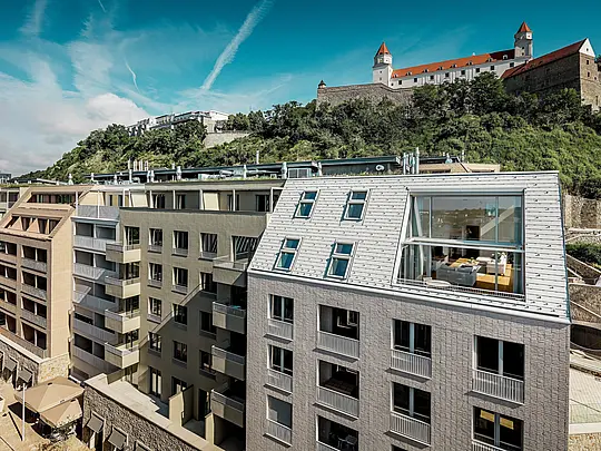 Sodobna strešna površina v mestni četrti Vydrica v Bratislavi s pogledom na grajski kompleks: strme strešne površine z velikimi okenskimi odprtinami in jasnimi linijami so elegantno poudarjene s strešnimi skodlami PREFA iz aluminija v barvah prefa bela in peščeno rjava. Natančna izdelava in harmonična povezava strehe in fasade ustvarjata močan vizualni poudarek nad strešno linijo mesta. PREFA aluminij prepriča s svojo lahkotnostjo, visoko odpornostjo proti vremenskim vplivom in trajno kakovostjo – idealno za kompleksne, arhitekturno zahtevne stanovanjske objekte.