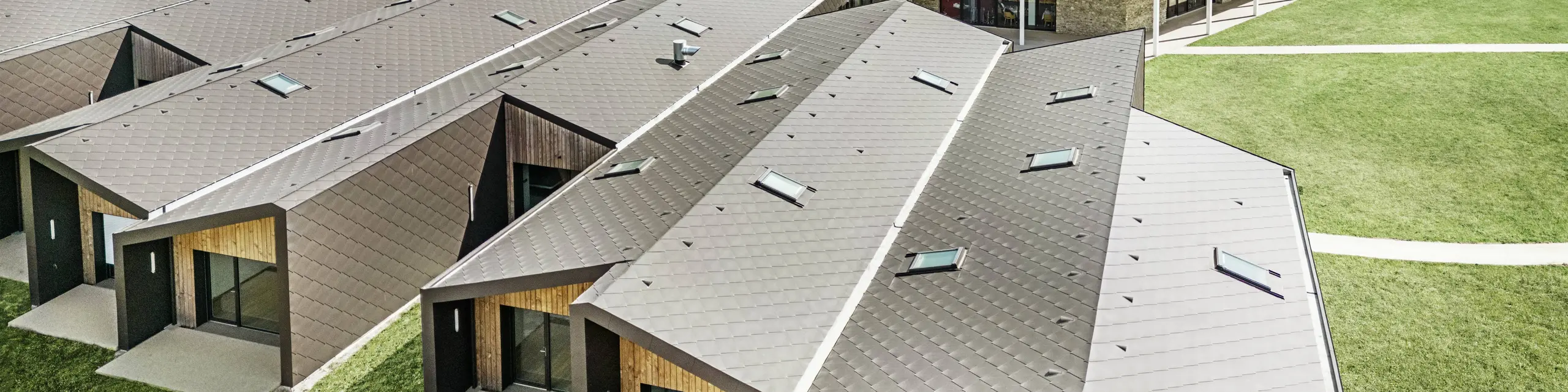 Slika prikazuje pogled iz zraka na sodoben stavbni kompleks, obložen s PREFA rombi 44 × 44 v barvi testa di moro. Za strešno površino so značilne geometrijske oblike v kombinaciji z velikimi steklenimi površinami in lesenimi elementi. V ozadju je vidna stara cerkev, ki tvori harmonično nasprotje sodobni arhitekturi. Zelena trata in okoliška narava poudarjata odprto in trajnostno zasnovo celotnega kompleksa.