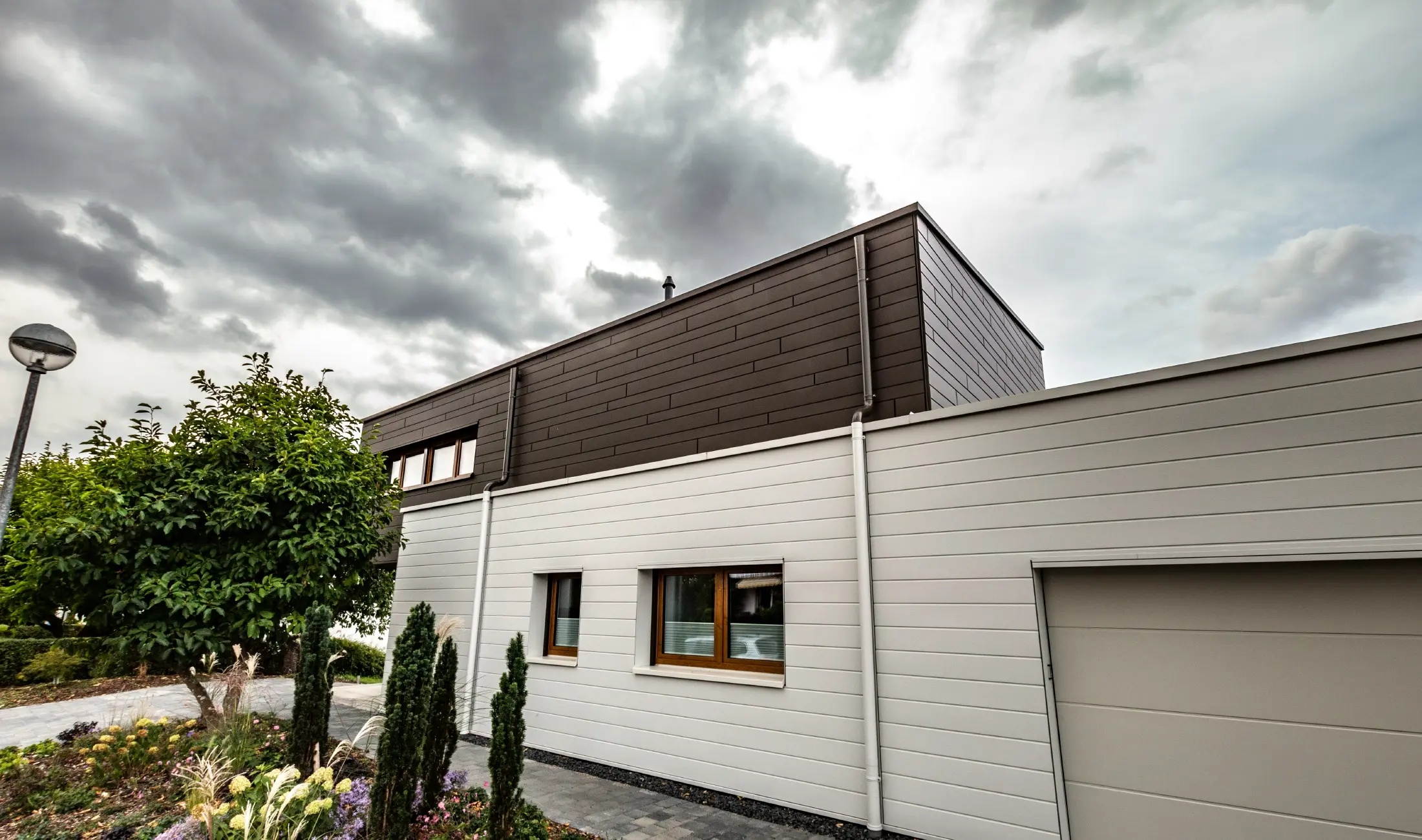 Letalski posnetek dostopa do moderne enodružinske hiše v Wippenhamu v Avstriji z dvobarvno PREFA aluminijasto fasado v bronasti in antracitni barvi. Hiša prikazuje stilsko integracijo fasadnih panelov Siding.X in lesene obloge. Jasno vidna je pokrita garaža, ki je brezhibno integrirana v arhitekturo hiše. Urejena vrt in terasa poudarjata sodoben in funkcionalen dizajn stavbe.