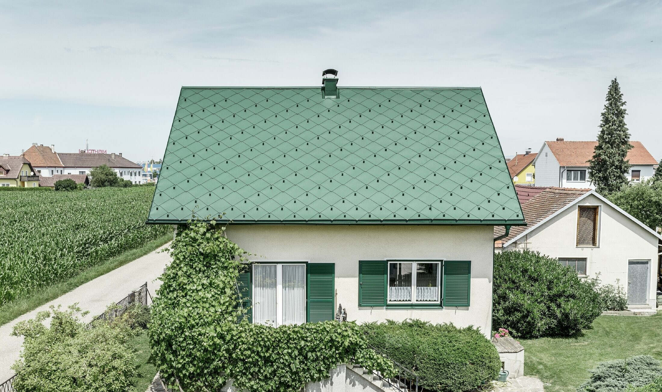 Klasična enodružinska hiša z dvokapno streho s strešno kritino iz aluminija PREFA v mahovo zeleni barvi z zelenimi polkni. Streha je bila izdelana s strešnimi rombi 44 × 44 PREFA v barvi P.10 mahovo zelena.