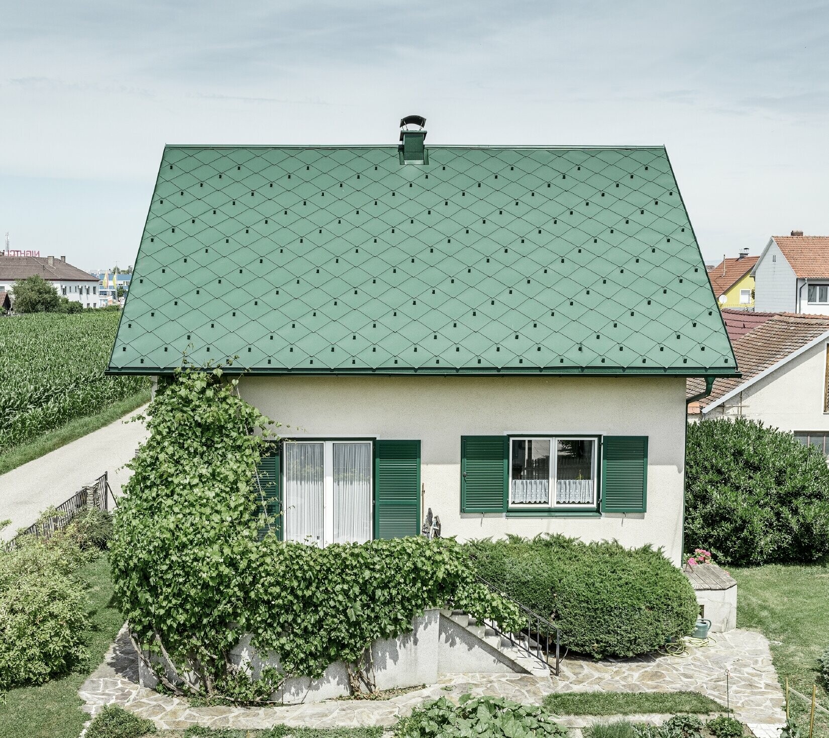 Klasična enodružinska hiša z dvokapno streho s strešno kritino iz aluminija PREFA v mahovo zeleni barvi z zelenimi polkni. Streha je bila izdelana s strešnimi rombi 44 × 44 PREFA v barvi P.10 mahovo zelena.
