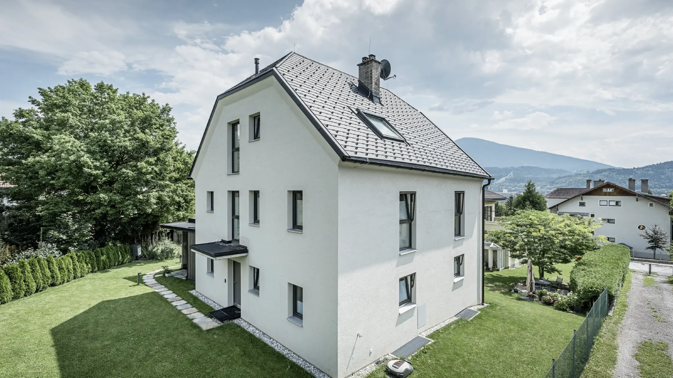 Moderna trietažna enodružinska hiša s svetlo fasado iz ometa in temnosivo dvokapno streho iz aluminija PREFA. Stavba ima več ozkih oken in odvodnjavanje strehe v antracitni barvi. Stoji v urejenem vrtu s travnikom, drevesi in živo mejo, v ozadju pa so vidne še druge stanovanjske hiše in gorska pokrajina.