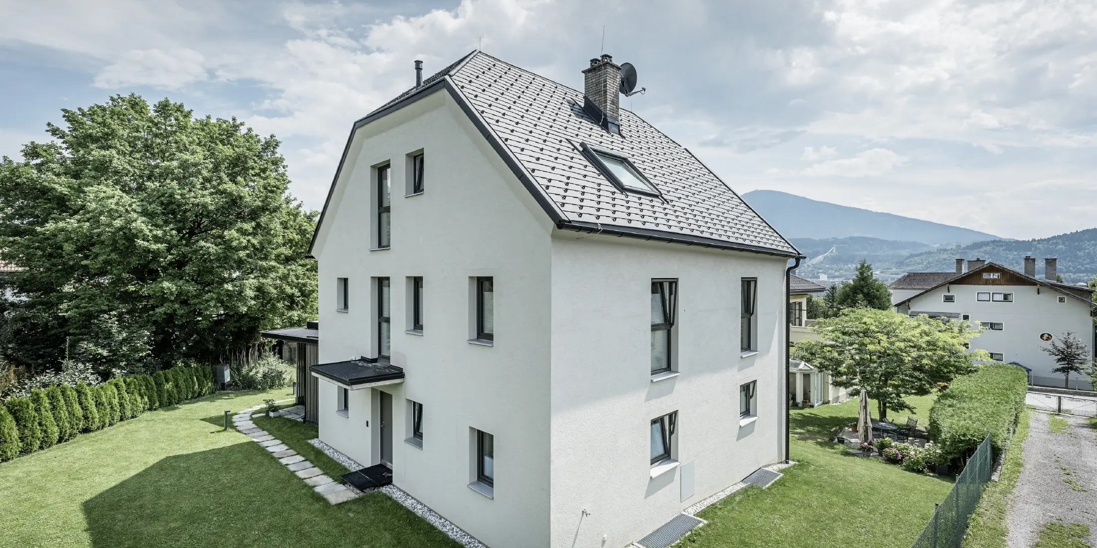 Moderna trietažna enodružinska hiša s svetlo fasado iz ometa in temnosivo dvokapno streho iz aluminija PREFA. Stavba ima več ozkih oken in odvodnjavanje strehe v antracitni barvi. Stoji v urejenem vrtu s travnikom, drevesi in živo mejo, v ozadju pa so vidne še druge stanovanjske hiše in gorska pokrajina.