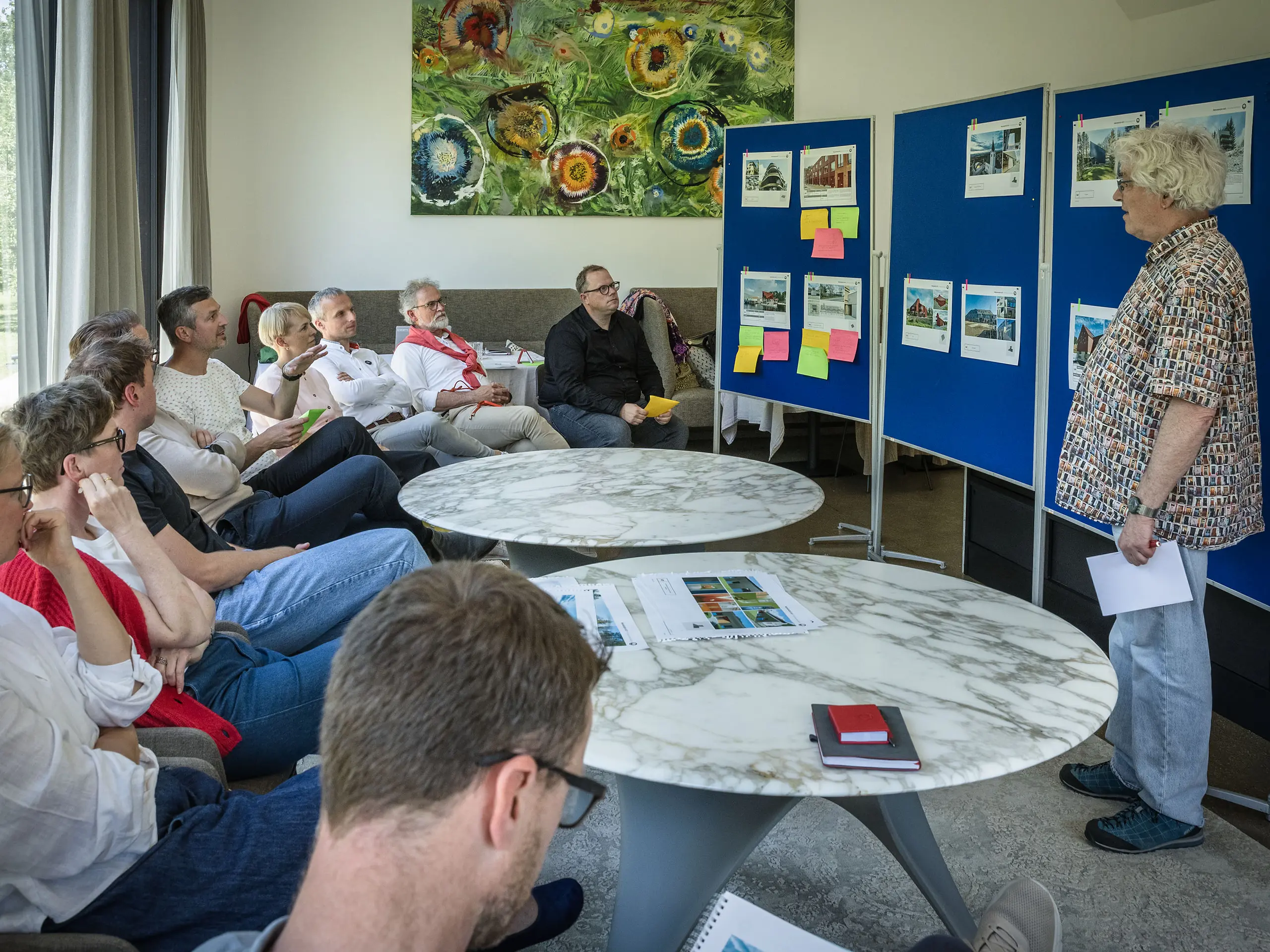 PREFARENZEN 2027 Zasedanje žirije v svetli seminarski dvorani: moderator Matthias Finkentey pred modrimi oglasnimi deskami predstavlja arhitekturne projekte iz PREFA aluminija, medtem ko žirija pozorno posluša in si dela zapiske. Velika okna prepuščajo dnevno svetlobo in ponujajo pogled na zeleni vrt. Na mizah ležijo knjige o arhitekturi, beležke in svinčniki. Prizor izraža zbrano analizo, ustvarjalno izmenjavo in ocenjevanje oblikovanja ter funkcionalnosti. PREFA aluminijasti izdelki simbolizirajo trajne, inovativne strehe in fasade, ki združujejo estetiko, kakovost in navdih za arhitekturne projekte.