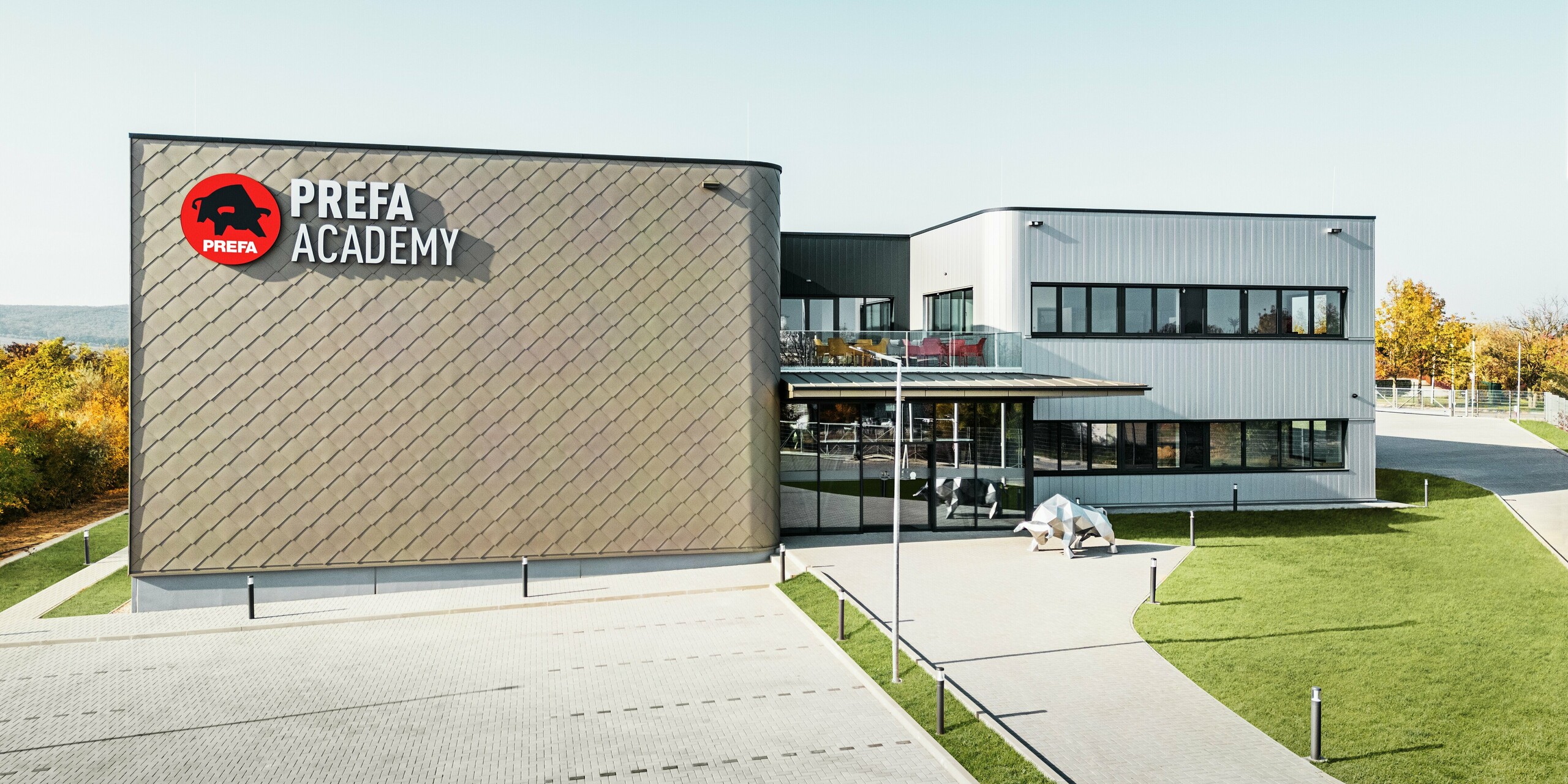 Nova lokacija PREFA Madžarska v Törökbálintu navdušuje s sodobno fasado iz aluminijastih rombov PREFA 44 × 44 v bronasti barvi ter fasadnih elementov PREFA Siding v srebrno kovinski barvi. Trpežni aluminijasti izdelki zagotavljajo vremensko odporno, enostavno za vzdrževanje in vizualno privlačno zunanjo podobo stavbe. Prepoznaven logotip PREFA in aluminijasta skulptura bika poudarjata inovativnost, kakovost in moč podjetja.