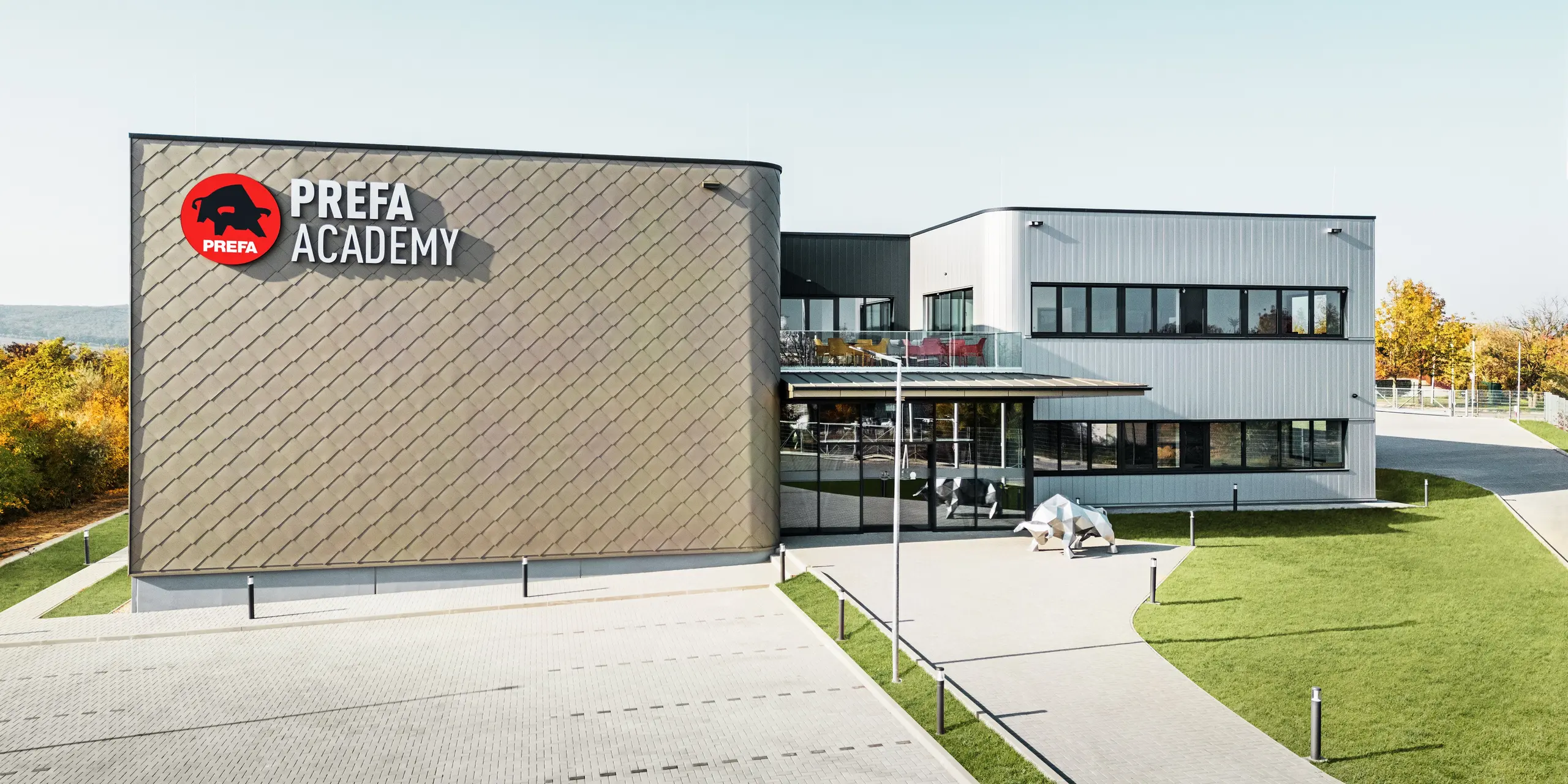 Nova lokacija PREFA Madžarska v Törökbálintu navdušuje s sodobno fasado iz aluminijastih rombov PREFA 44 × 44 v bronasti barvi ter fasadnih elementov PREFA Siding v srebrno kovinski barvi. Trpežni aluminijasti izdelki zagotavljajo vremensko odporno, enostavno za vzdrževanje in vizualno privlačno zunanjo podobo stavbe. Prepoznaven logotip PREFA in aluminijasta skulptura bika poudarjata inovativnost, kakovost in moč podjetja.