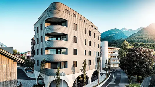 Stranski pogled na hišo Bernerhaus v Buchsu z značilno trikotno obliko in mehko zaobljenimi koti. Neprekinjena fasada iz PREFA fasadnih skodel v barvi P.10 svetlo siva obdaja stavbo kot luskasta aluminijasta koža in poudarja tekoče balkonske pasove. Nad masivnim betonskim podstavkom z elegantnimi arkadami deluje lahka, vremensko odporna PREFA aluminijasta fasada še posebej izrazito. Natančno položena, trajna in z minimalnim vzdrževanjem se harmonično vklaplja v alpsko okolje in daje močno arhitekturno sporočilo.