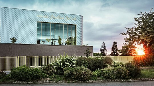  Moderna šolska stavba Collège Léontine Dolivet v Cesson-Sévigné z izrazito aluminijasto fasado iz okoli 15.000 PREFA fasadnih rombov v kovinsko srebrni barvi. Bleščeča površina odbija svetlobo in ustvarja živahen, eleganten videz. Velika steklena fasada odpira pogled na zelenje, robustna PREFA fasadna rešitev pa zagotavlja dolgo življenjsko dobo in odpornost proti vremenskim vplivom. Kombinacija spreminjajočega se kovinskega videza in jasne arhitekture naredi stavbo pravo pašo za oči in poudarja prednosti aluminija: lahkost, brez vzdrževanja in trajnost.