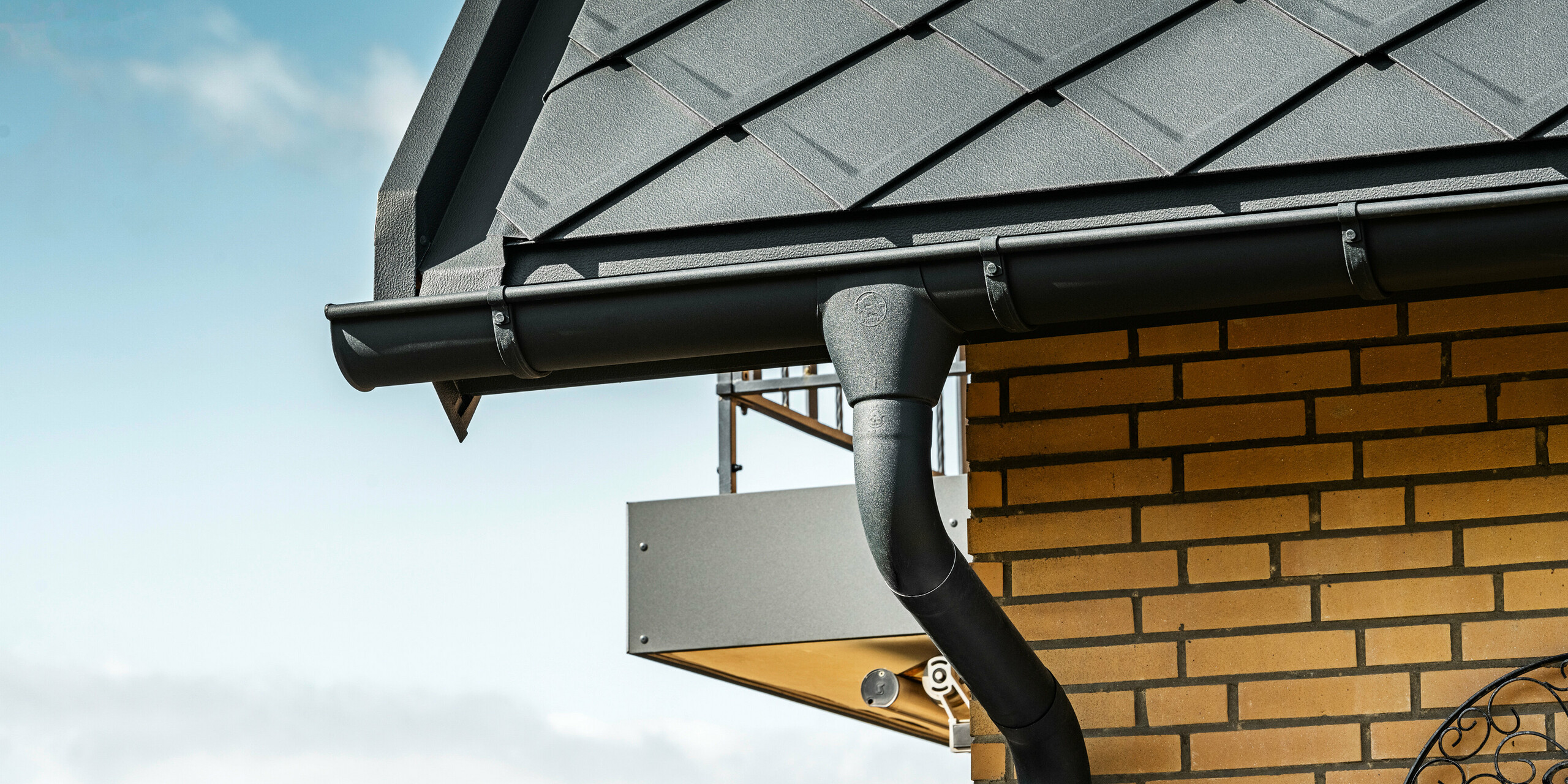 Podroben prikaz PREFA sistema za odvodnjavanje strehe enodružinske hiše s PREFA strešnimi rombi 29×29 antracitne barve, ki izstopajo na strehi v ozadju. Vidite lahko žlebove in odtočno cev v P.10 antracit barvi, ki se brezhibno prilegajo celotnemu dizajnu. Rumene opečne stene hiše dajejo naraven barvni kontrast sodobnemu aluminijastemu strešnemu sistemu.