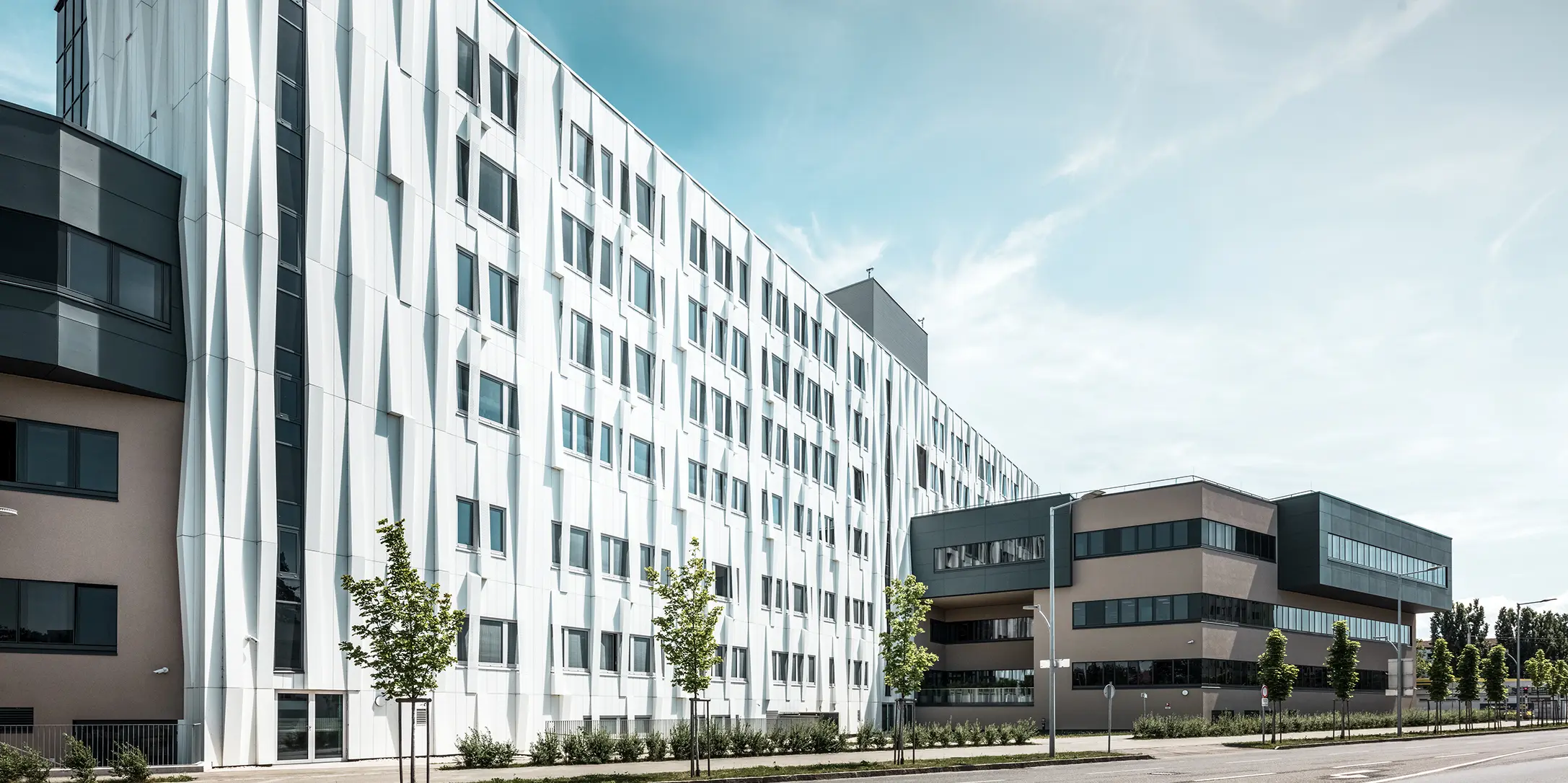 Stranski pogled na univerzitetno kliniko v St. Pöltenu, Spodnja Avstrija. Robustna aluminijasta fasada stavbo spreminja v pravo znamenitost mesta. V ospredju je vidna cesta z nekaj drevesi.