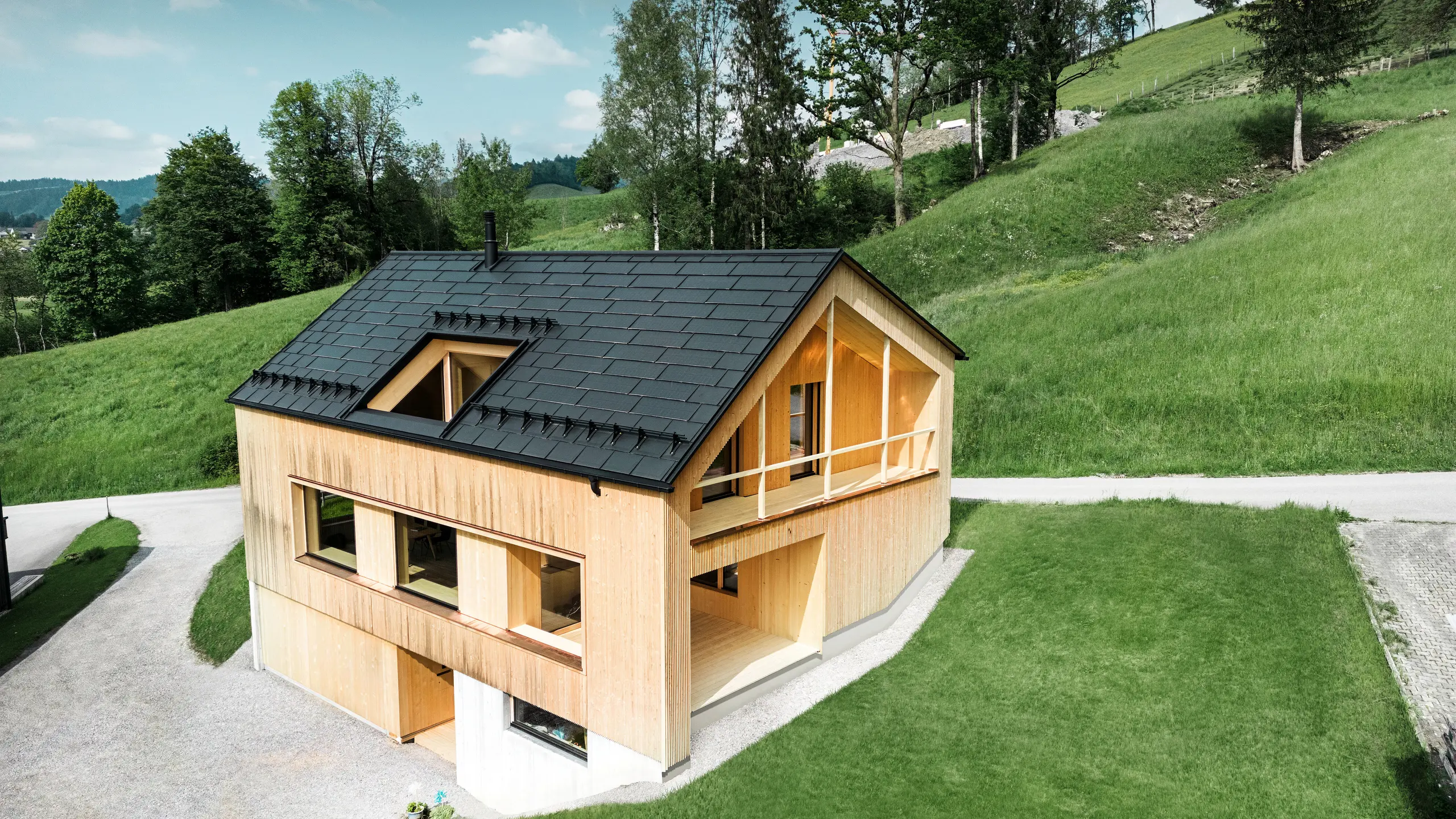 Slika prikazuje slikovito ekološko hišo v Eggu, obdano z zelenimi griči, s trajnostno solarno strešno ploščo PREFA. Črne, visokokakovostne PREFA strešne plošče R.16 in solarne strešne plošče PREFA se harmonično vklapljajo v podeželsko pokrajino in poudarjajo naravno lepoto okolice. Solarne strešne plošče niso le izraz zavezanosti obnovljivi energiji, ampak tudi oblikovni element, ki stilsko dopolnjuje moderno leseno fasado in čiste linije stavbe. Ta hiša je vzor za okolju prijazno gradnjo in navdih za prihodnostno usmerjeno arhitekturo.