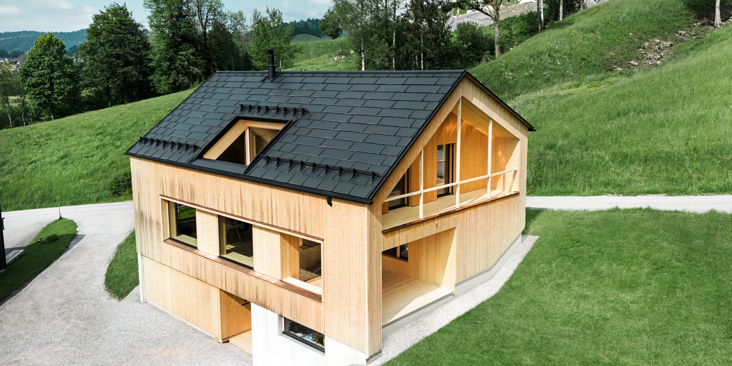 Slika prikazuje slikovito ekološko hišo v Eggu, obdano z zelenimi griči, s trajnostno solarno strešno ploščo PREFA. Črne, visokokakovostne PREFA strešne plošče R.16 in solarne strešne plošče PREFA se harmonično vklapljajo v podeželsko pokrajino in poudarjajo naravno lepoto okolice. Solarne strešne plošče niso le izraz zavezanosti obnovljivi energiji, ampak tudi oblikovni element, ki stilsko dopolnjuje moderno leseno fasado in čiste linije stavbe. Ta hiša je vzor za okolju prijazno gradnjo in navdih za prihodnostno usmerjeno arhitekturo.